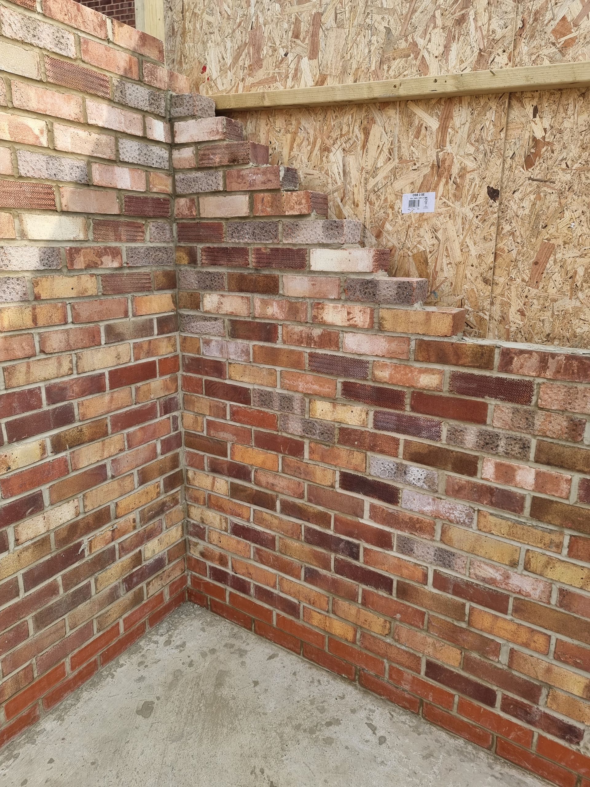 Brick corner with unfinished bricks meeting a wood-paneled wall, set on a concrete floor.