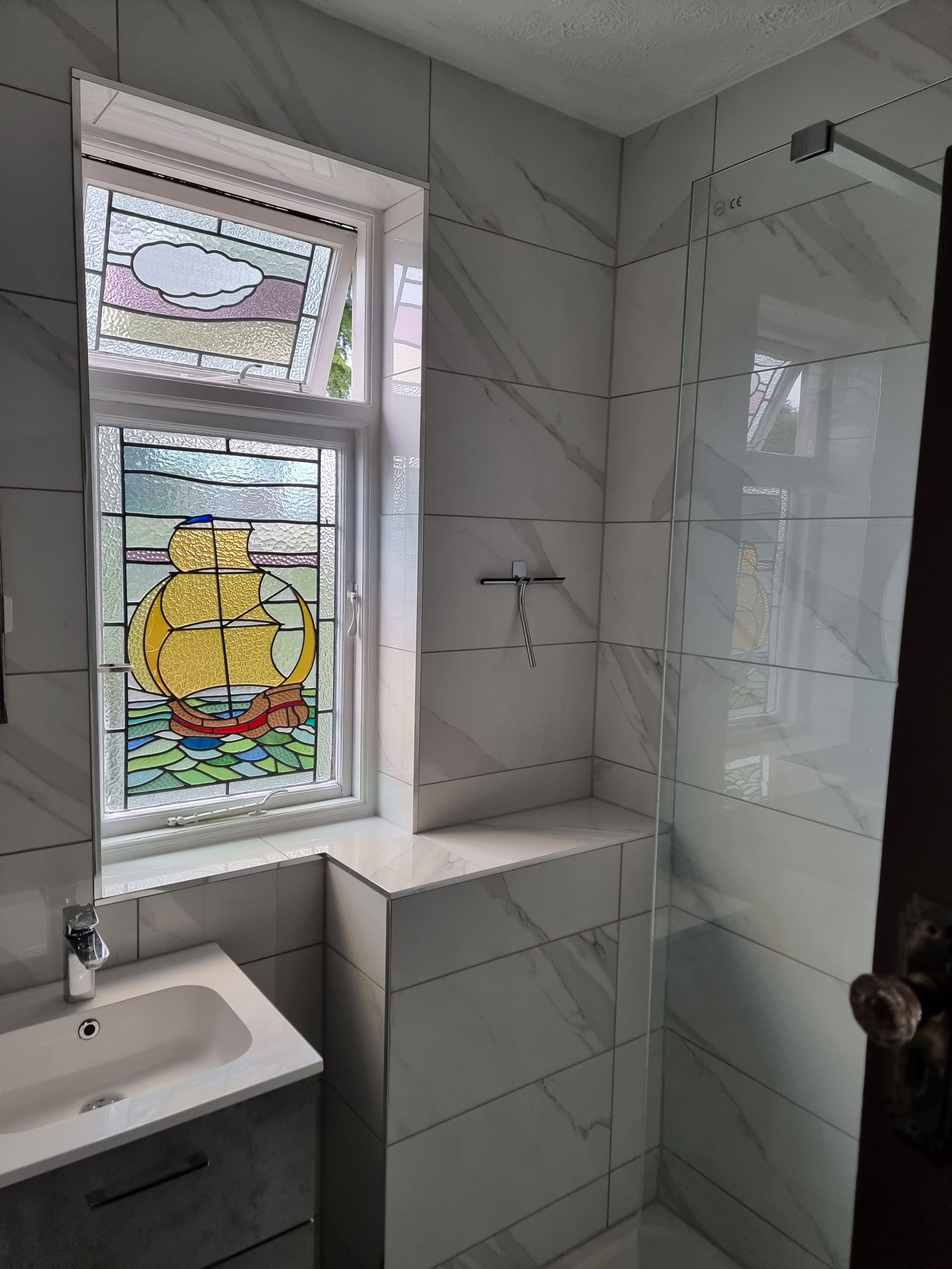 Bathroom with a stained-glass window depicting a ship. White marble-like tiles and glass shower door. Sink on the left.