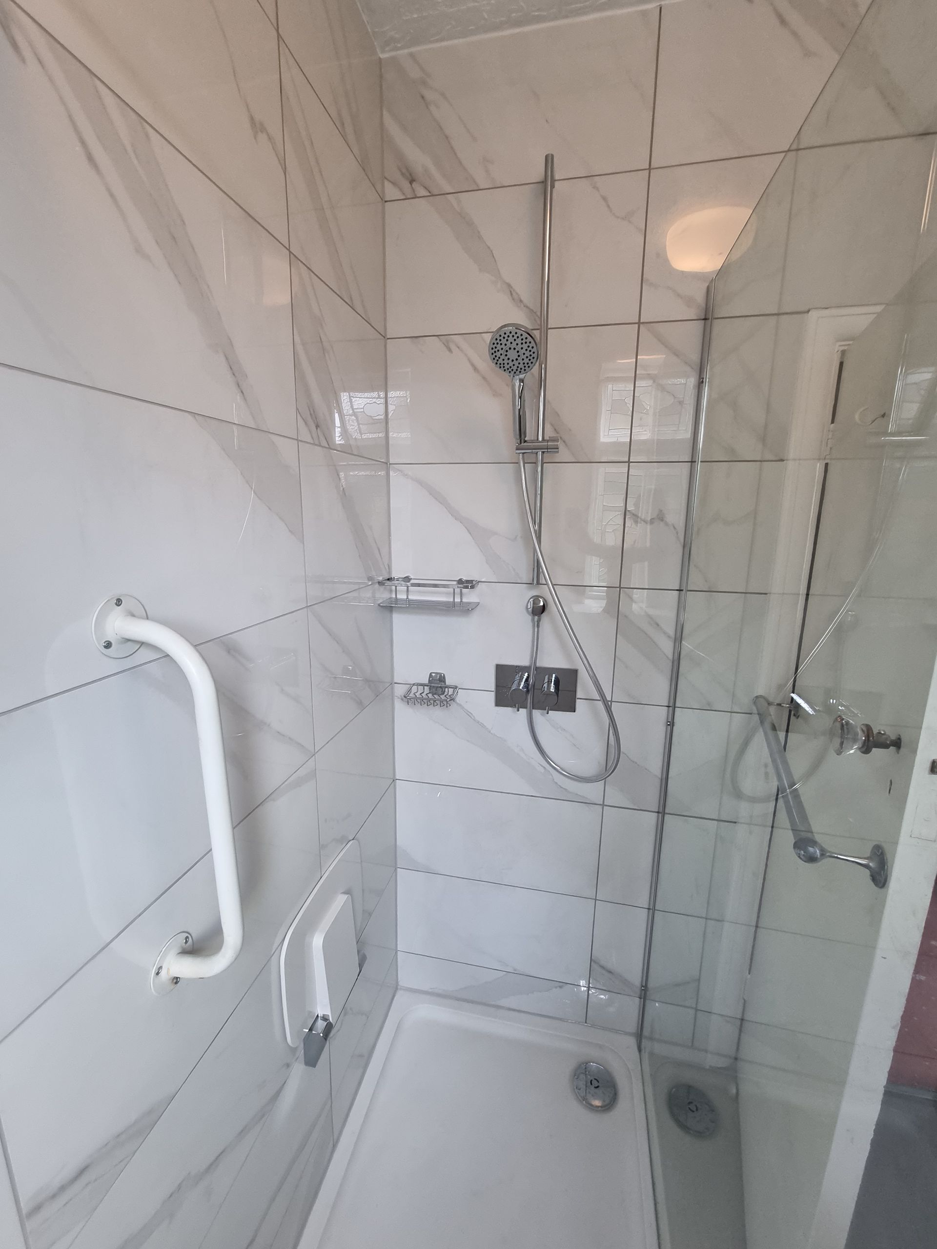 White tile shower stall with grab bars, showerhead, and glass door.