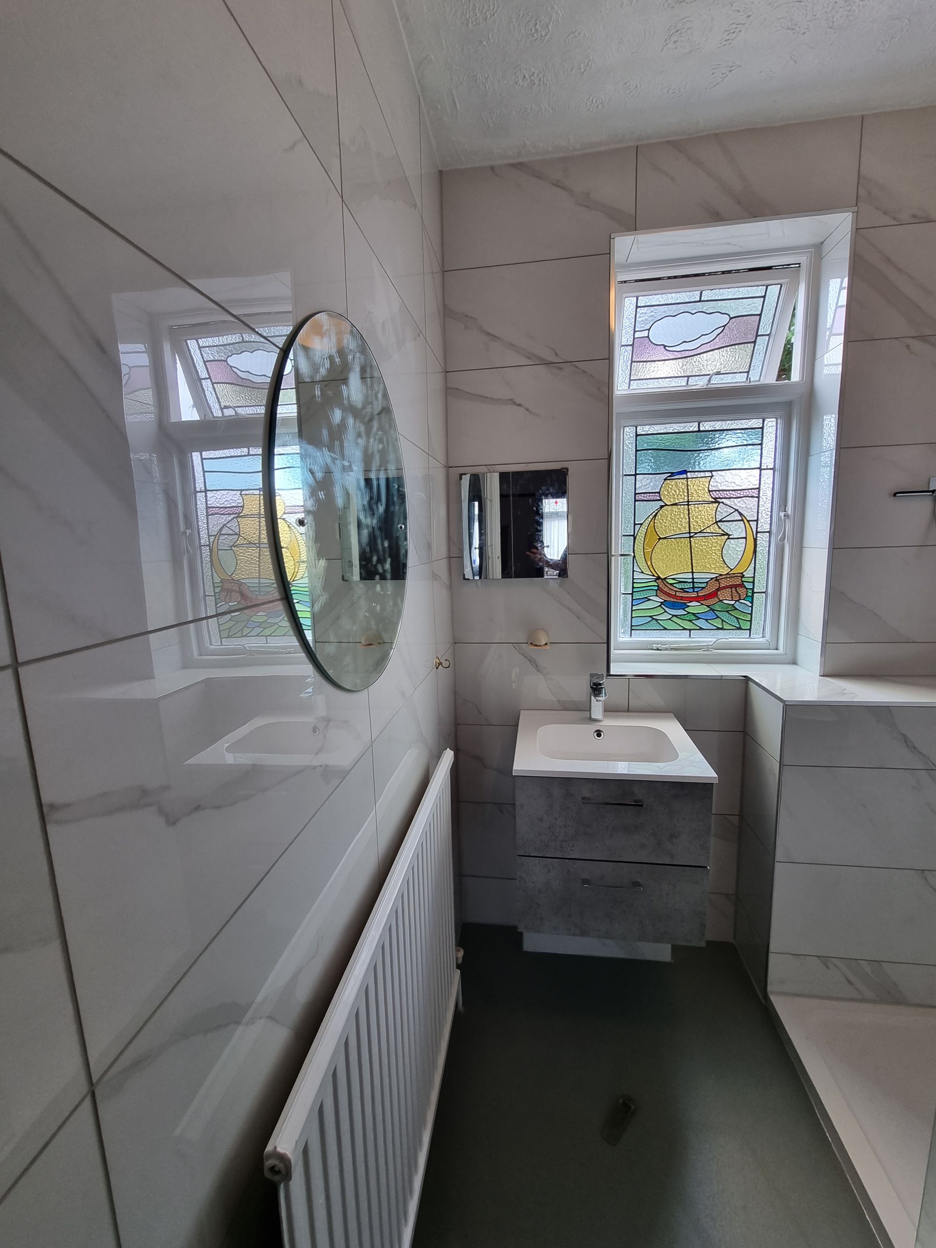 Bathroom with white marble-look tile, a sink, and a stained glass window. A round mirror hangs on the wall.
