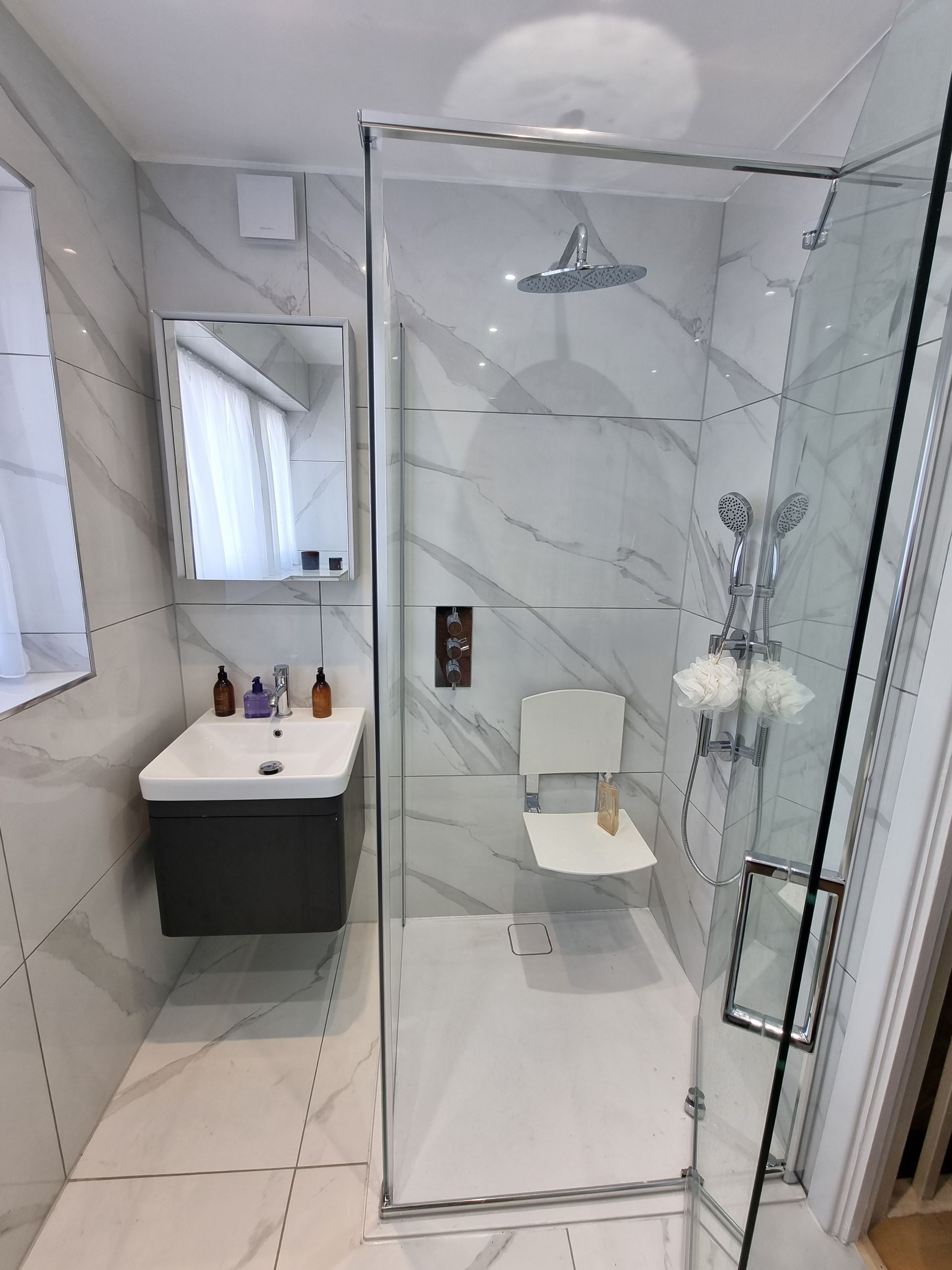 Accessible bathroom with shower, sink, and safety seat; white and gray marble tiles.