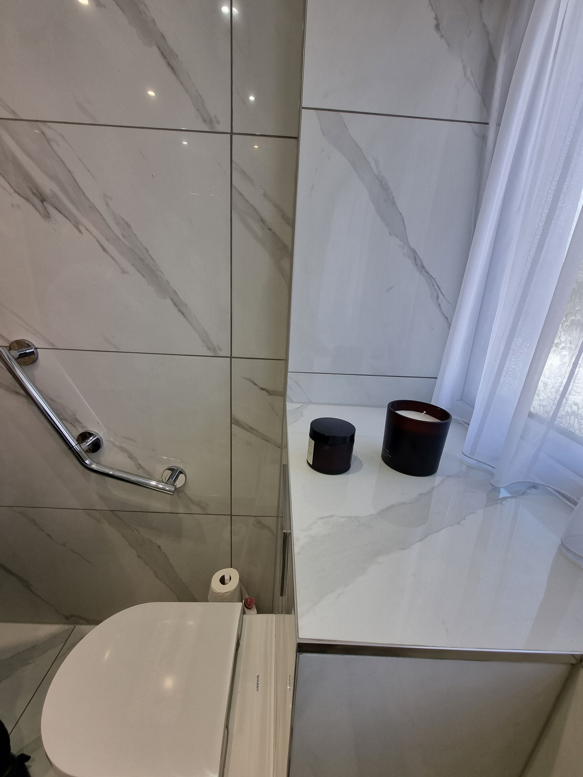 White marble-tiled bathroom with a toilet, grab bar, and candles on a ledge.