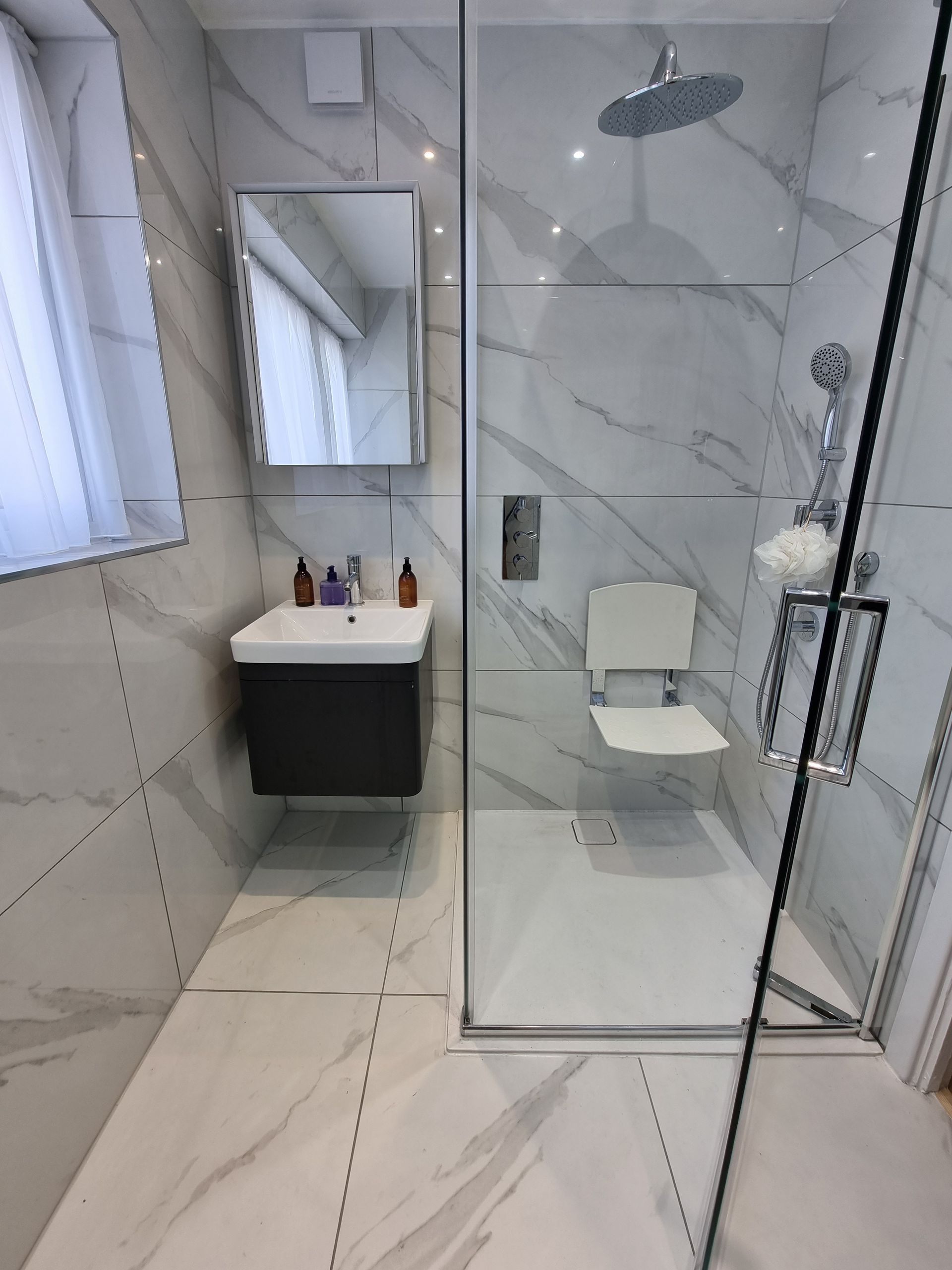Bathroom with marble-look tile, glass shower, wall-mounted sink, and accessible shower seat.