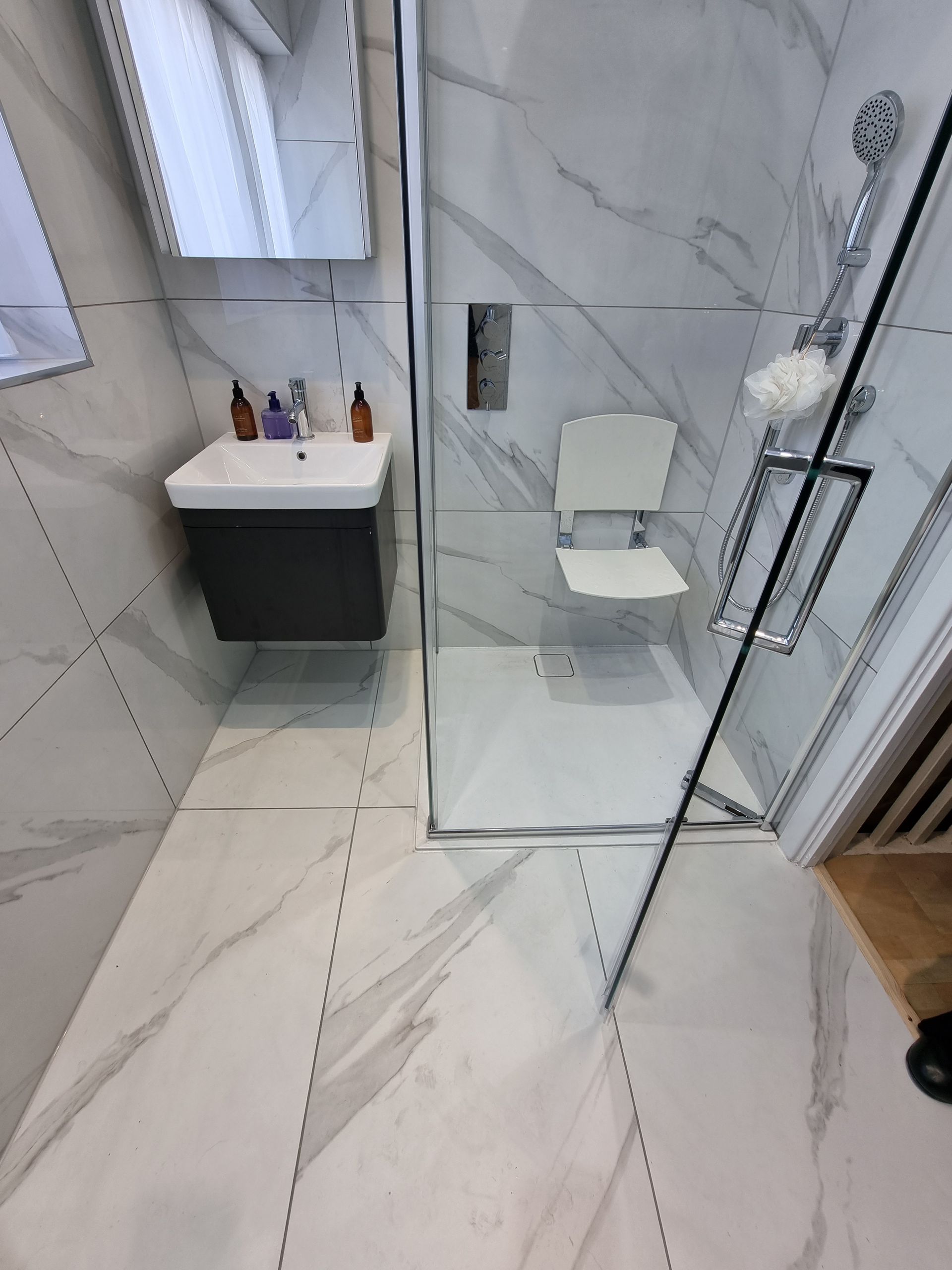 Modern bathroom with accessible shower and floating vanity. White marble tile, shower chair, silver fixtures.
