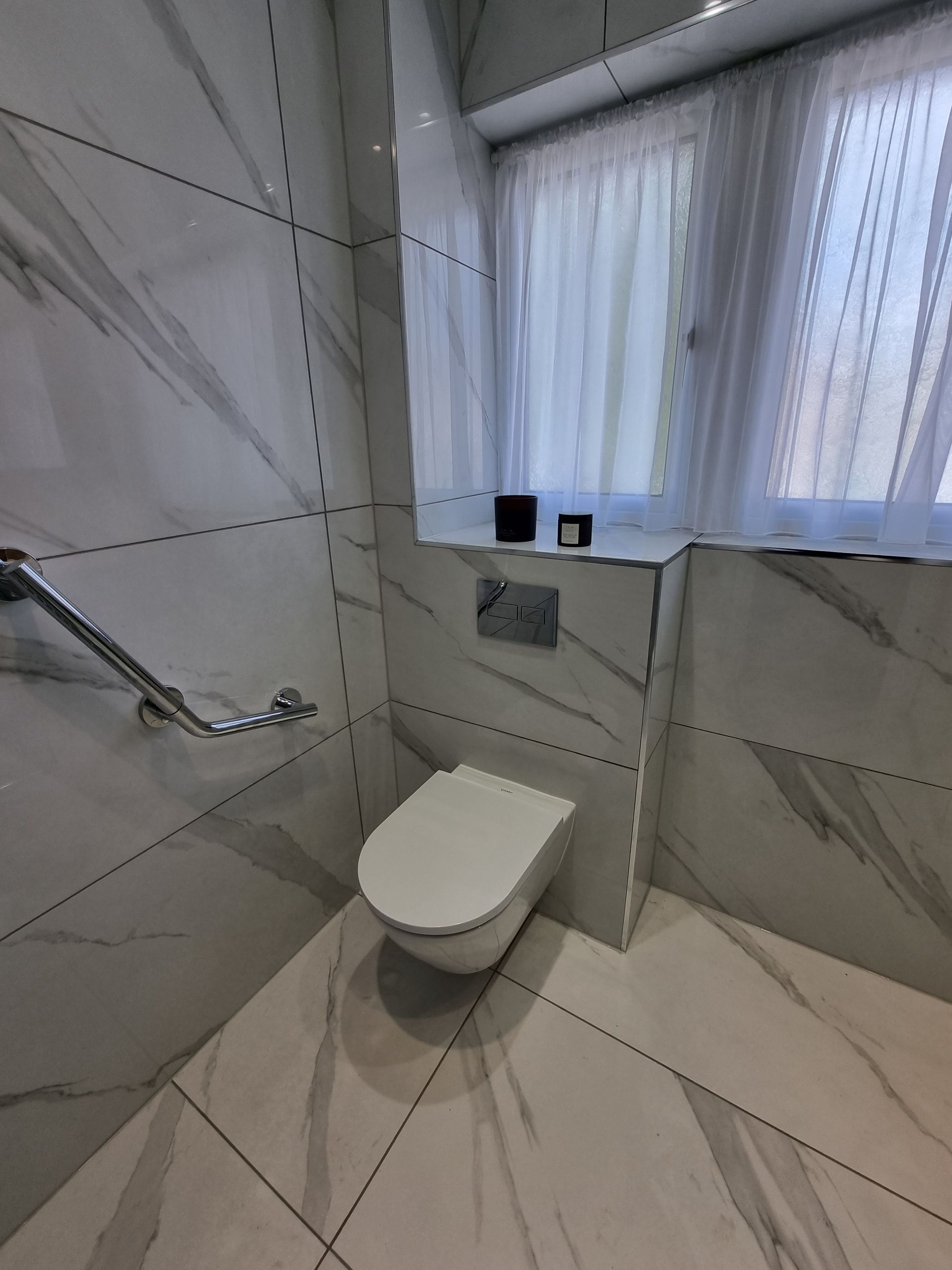 White tiled accessible bathroom with wall-mounted toilet, grab bar, and window with sheer curtains.