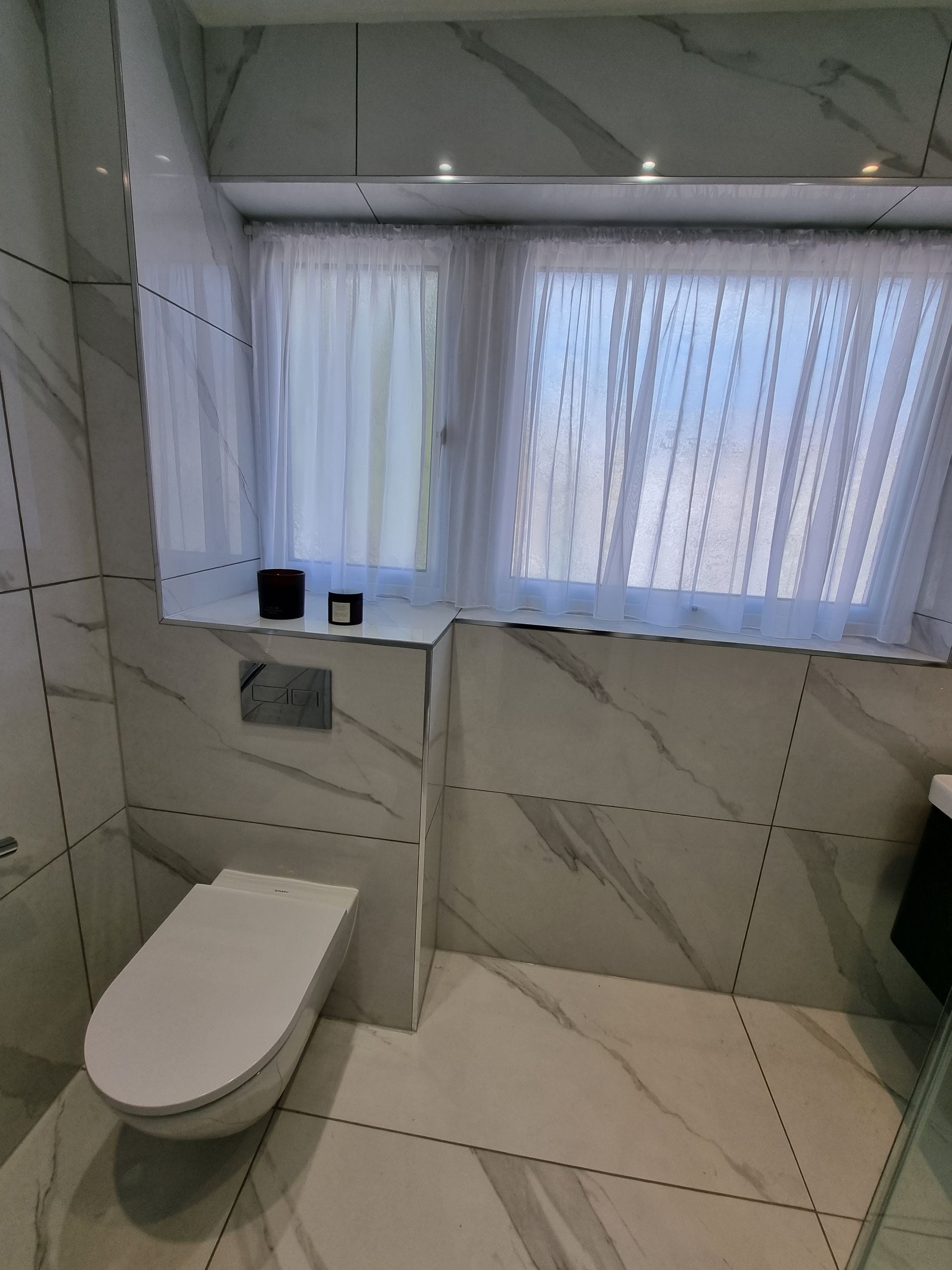 Modern bathroom with white marble tile, toilet, and window with sheer curtains.