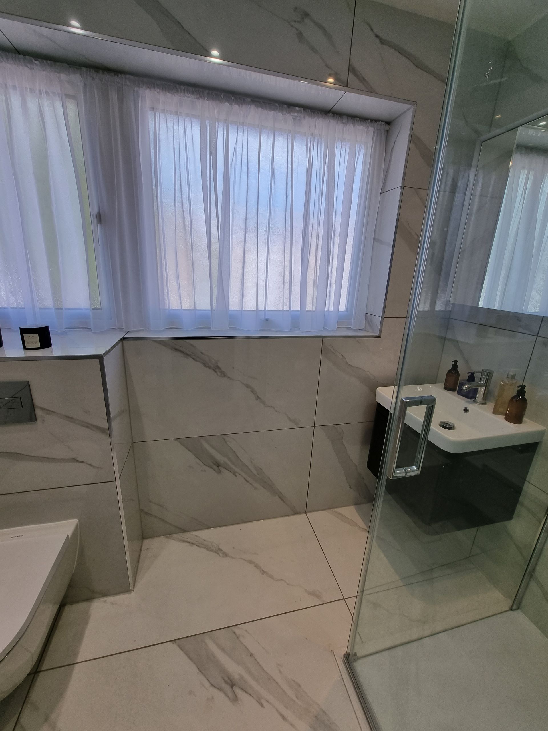 Bathroom with marble-look tiles, a window with sheer curtains, a wall-mounted sink, and a glass shower enclosure.