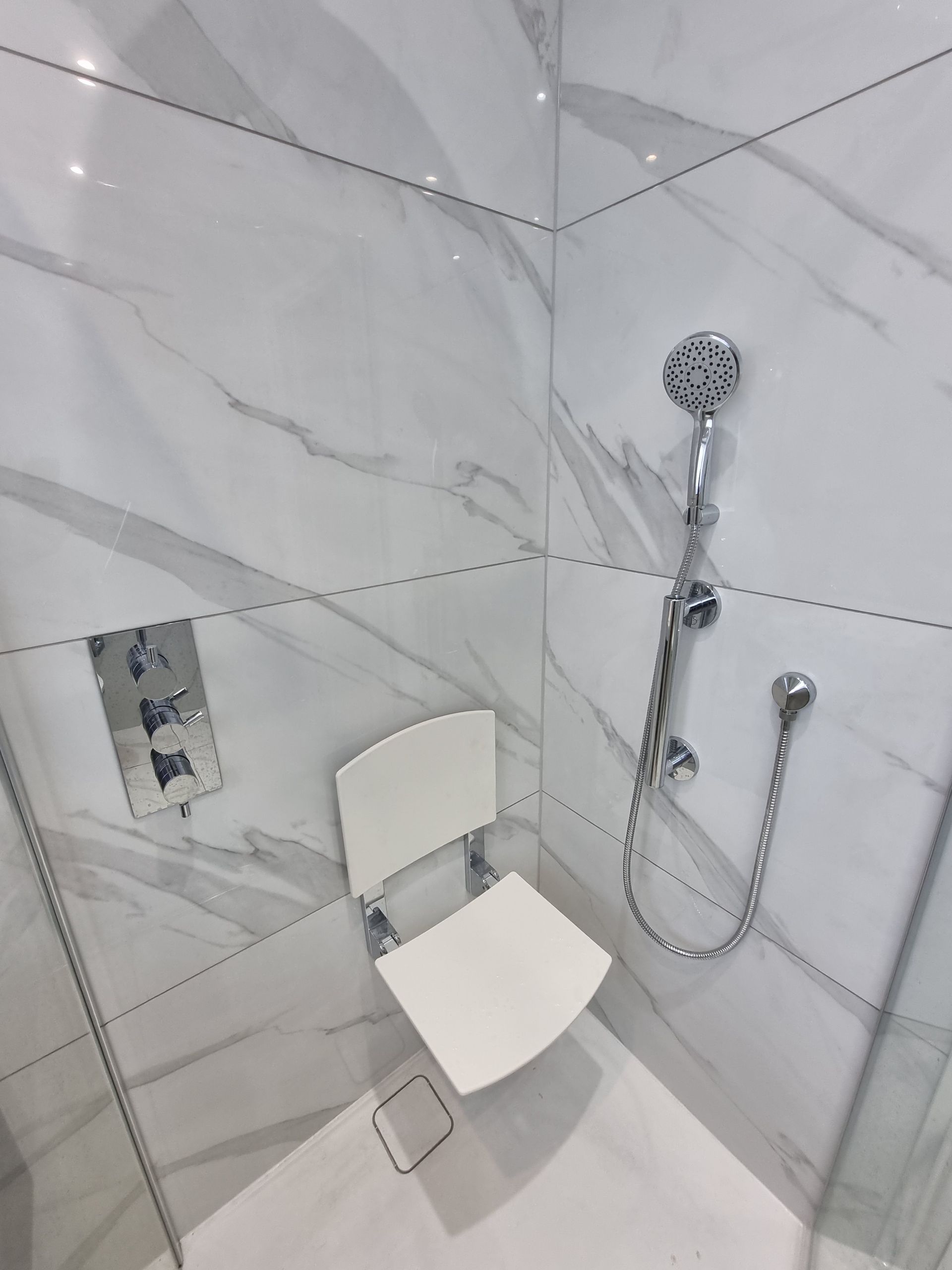 Shower with marble-look tile, hand shower, and a fold-down white seat for accessibility.