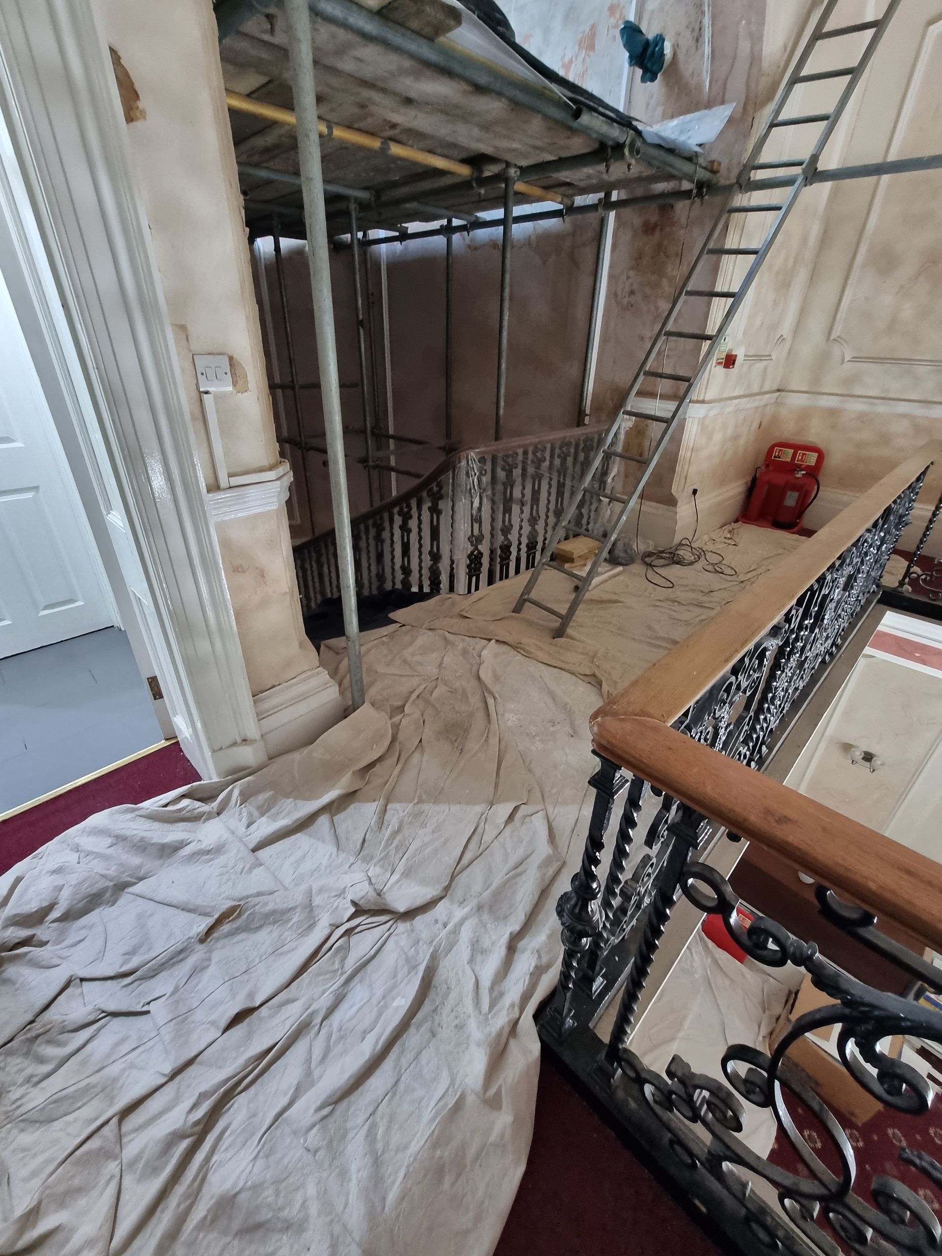 Staircase under renovation with scaffolding, ladder, and protective coverings; some paintwork is visible.