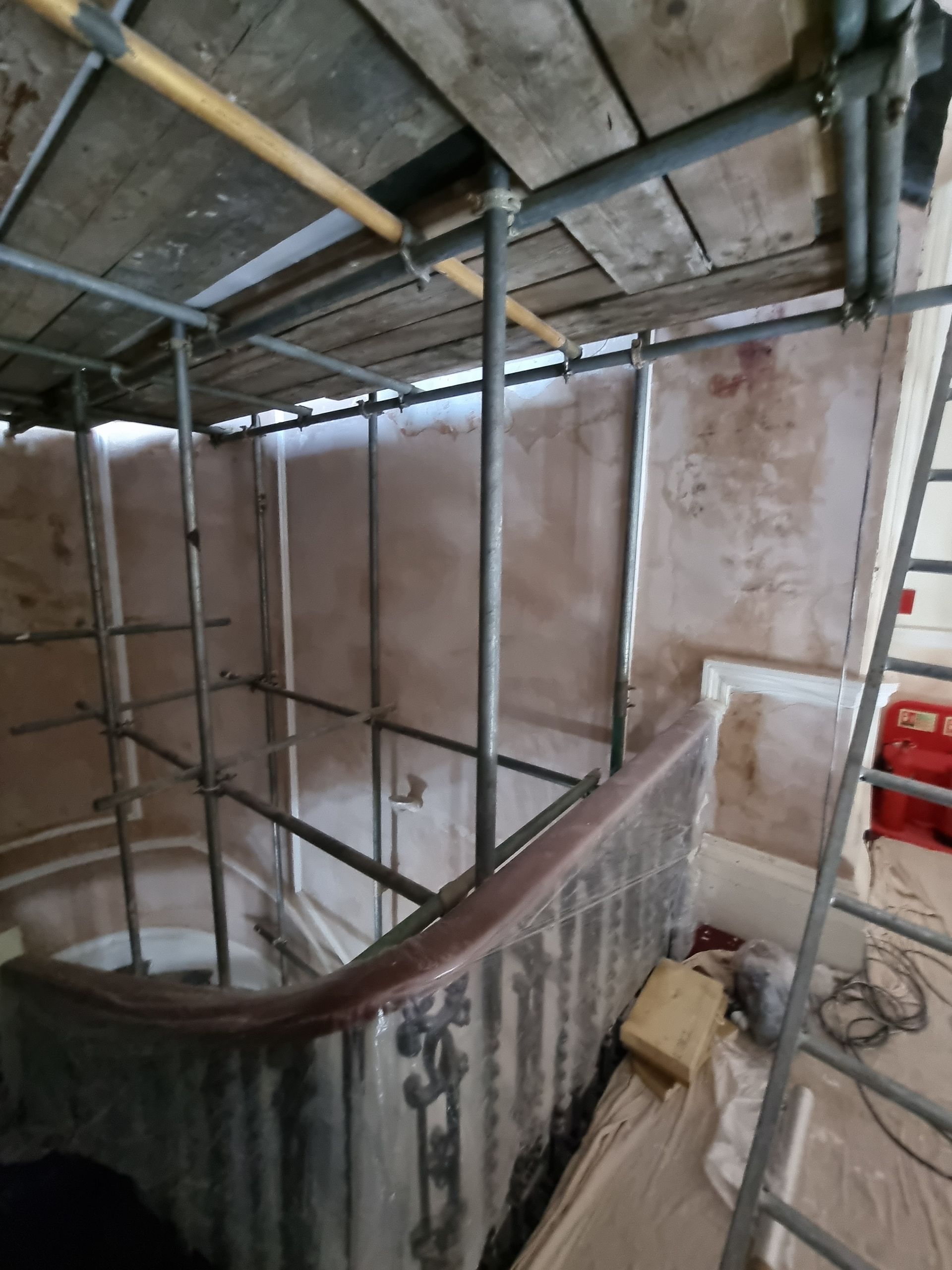 Interior view of scaffolding around a curved staircase. Walls are textured plaster. Brown wood handrail.