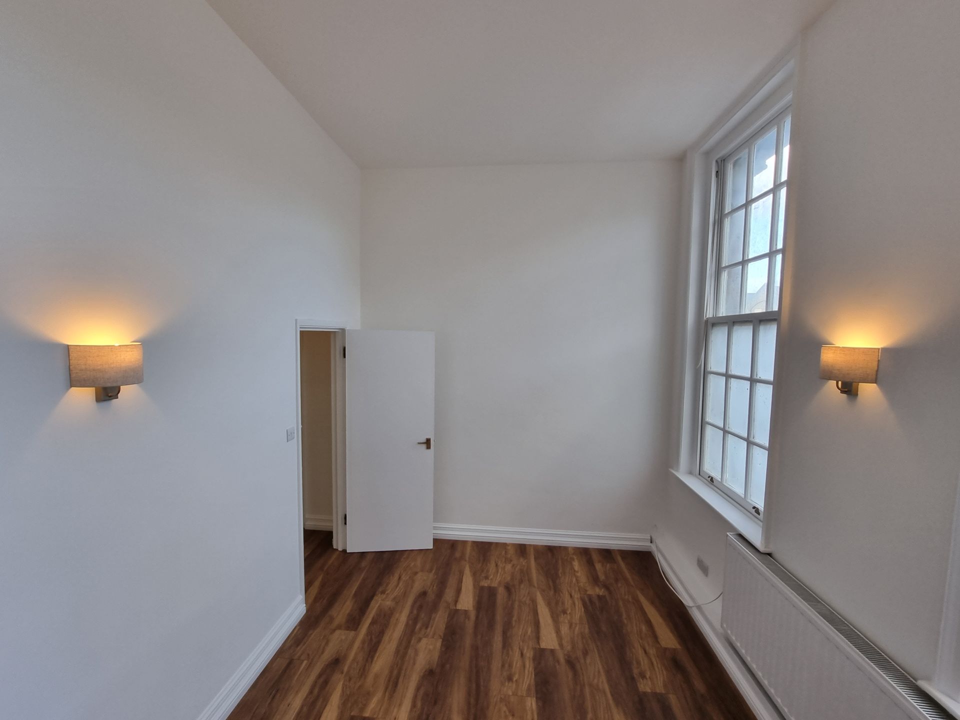 Empty room with white walls, wood floor, open door, large window with radiator, and sconces.