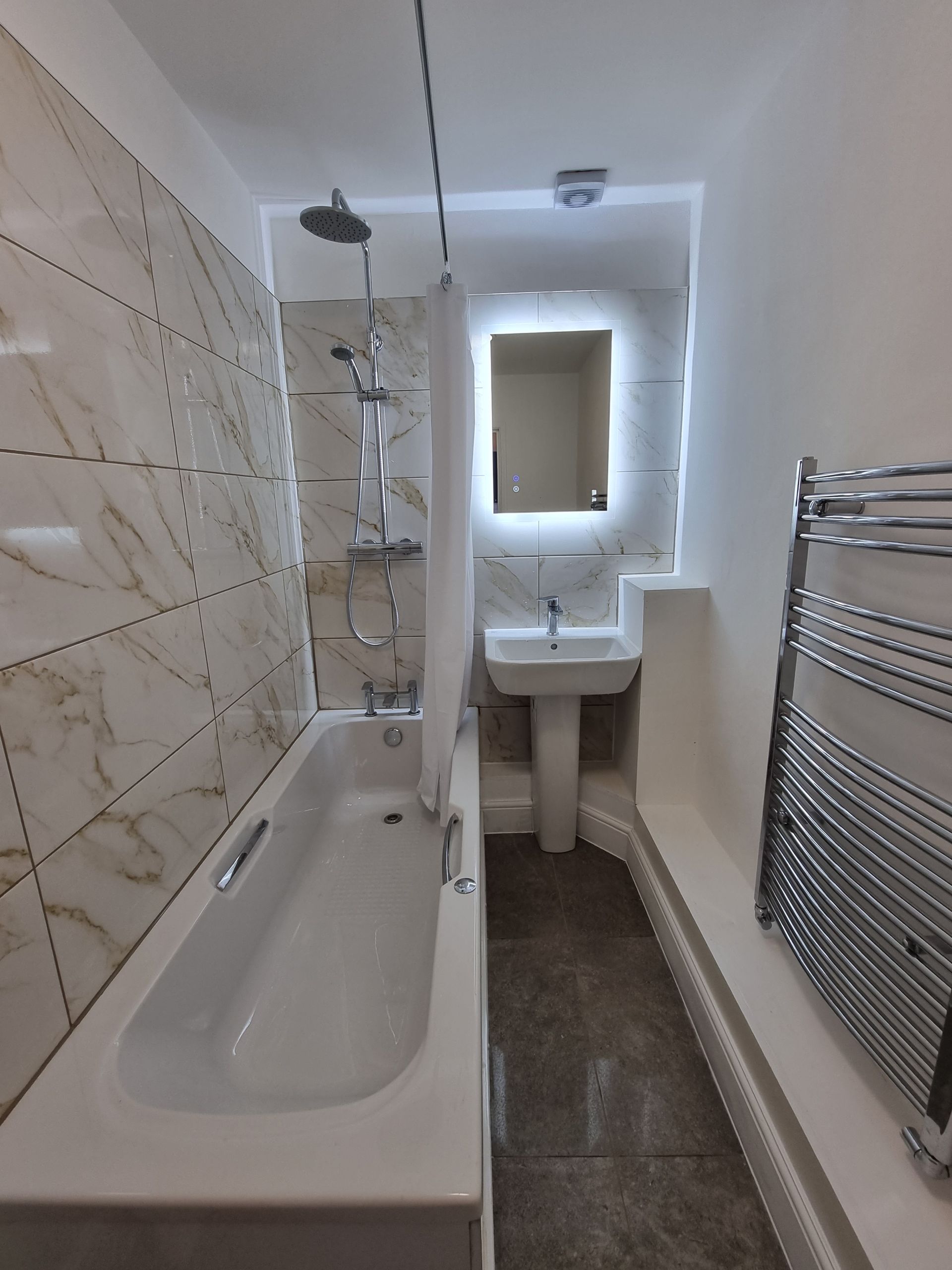 Narrow bathroom with white tub, shower, sink, and heated towel rack. Marble-patterned tile on one wall.