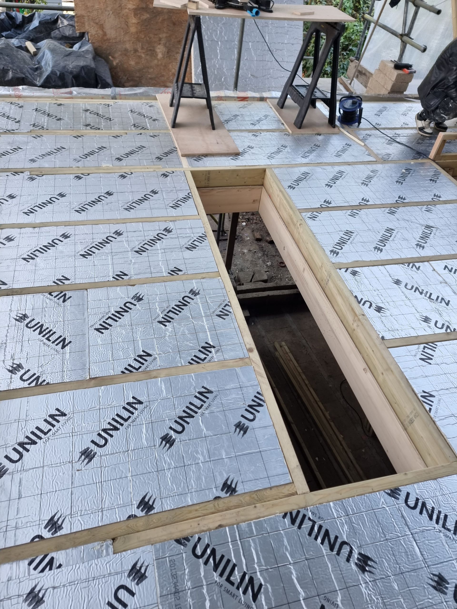 Wooden floor under construction; insulation sheets visible between beams, opening in floor.
