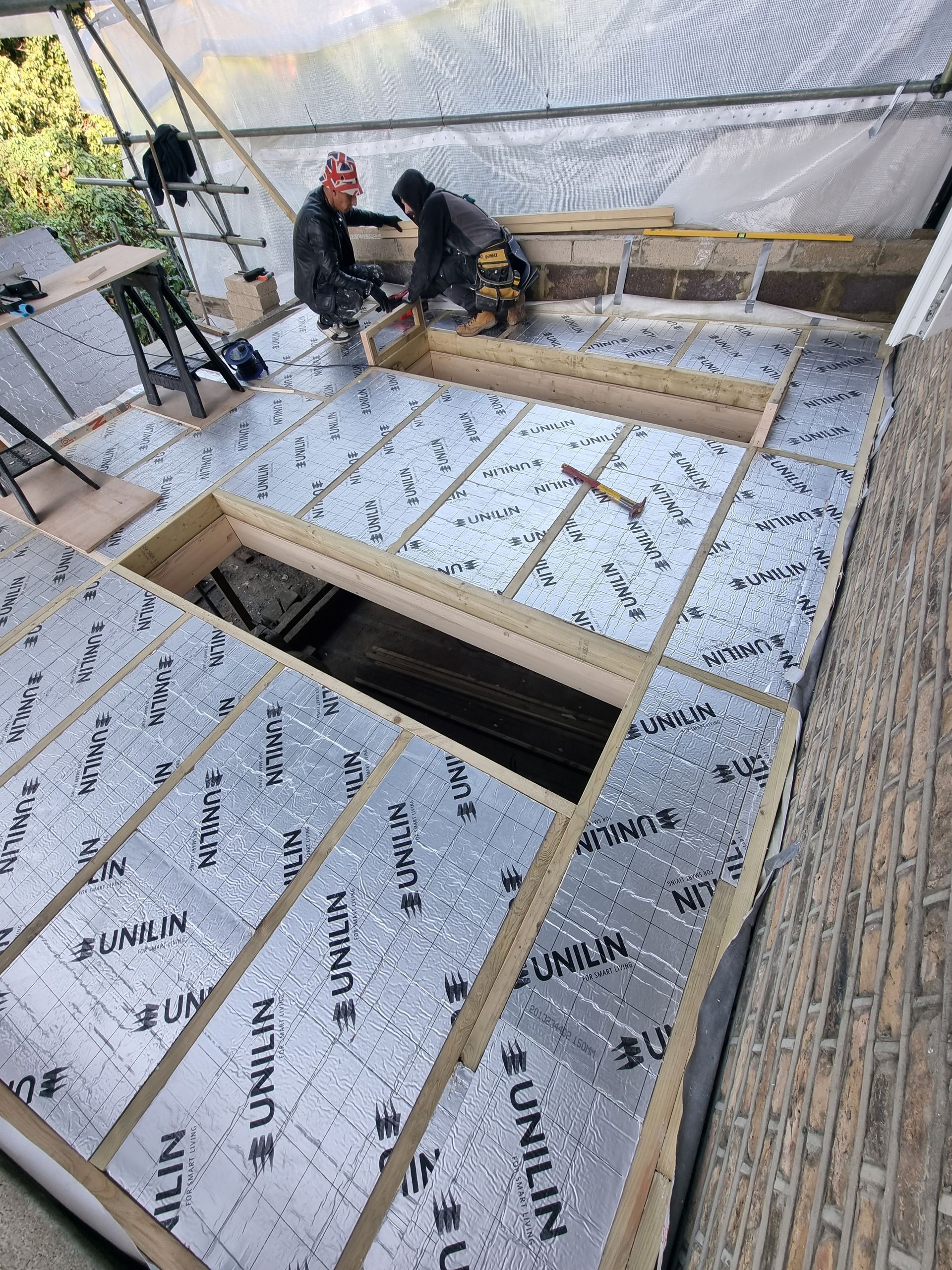 Two workers installing insulation on a roof. Construction site; silver insulation, wood beams.