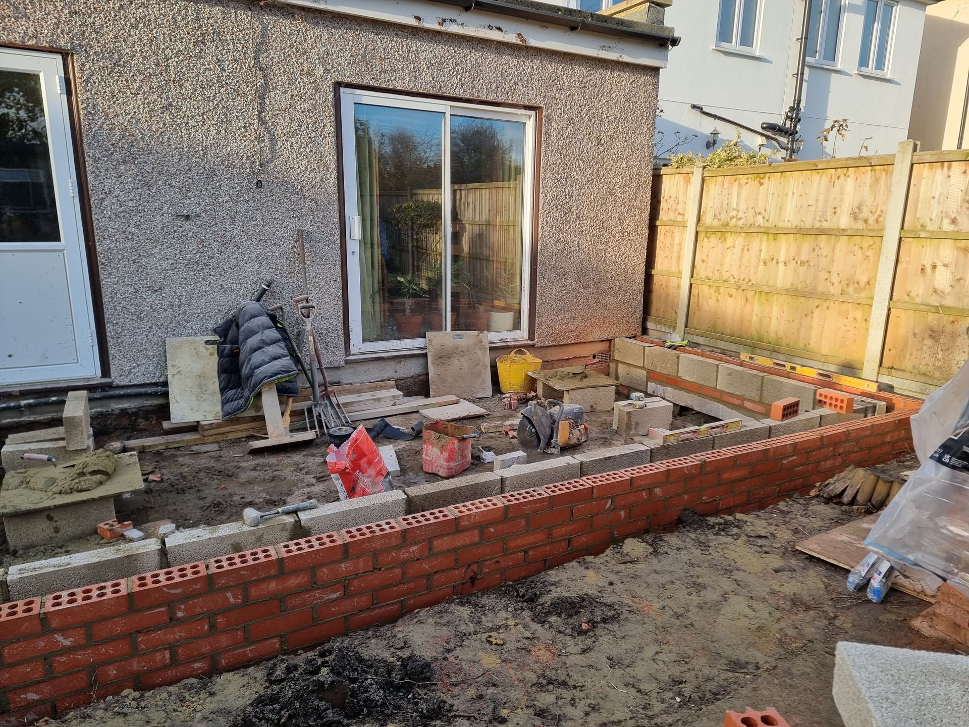 Bricklaying in progress on a garden patio next to a house with a sliding glass door and a wooden fence.