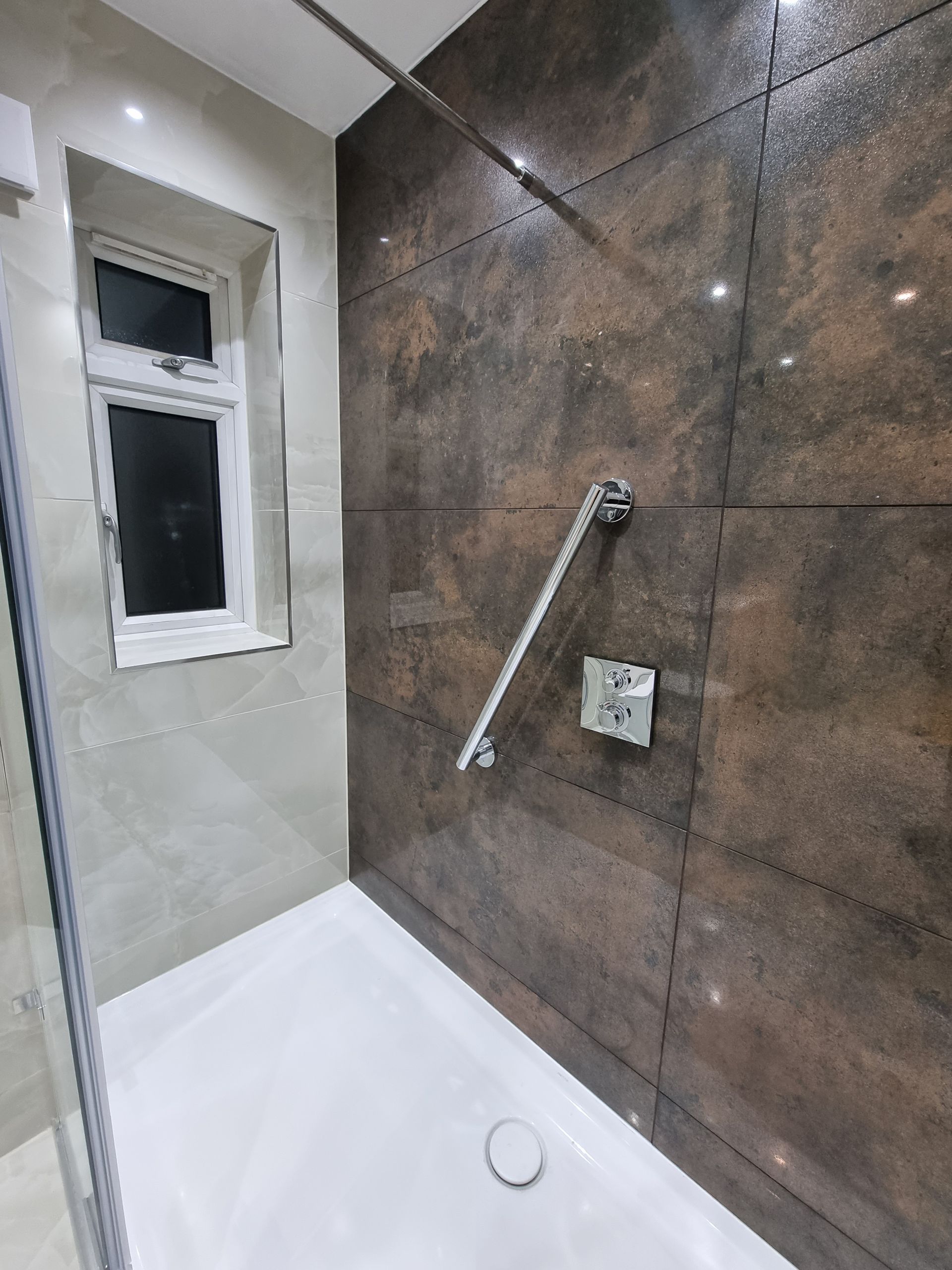 Shower stall with brown tiled wall, white shower base, and window. Includes a grab bar and shower controls.