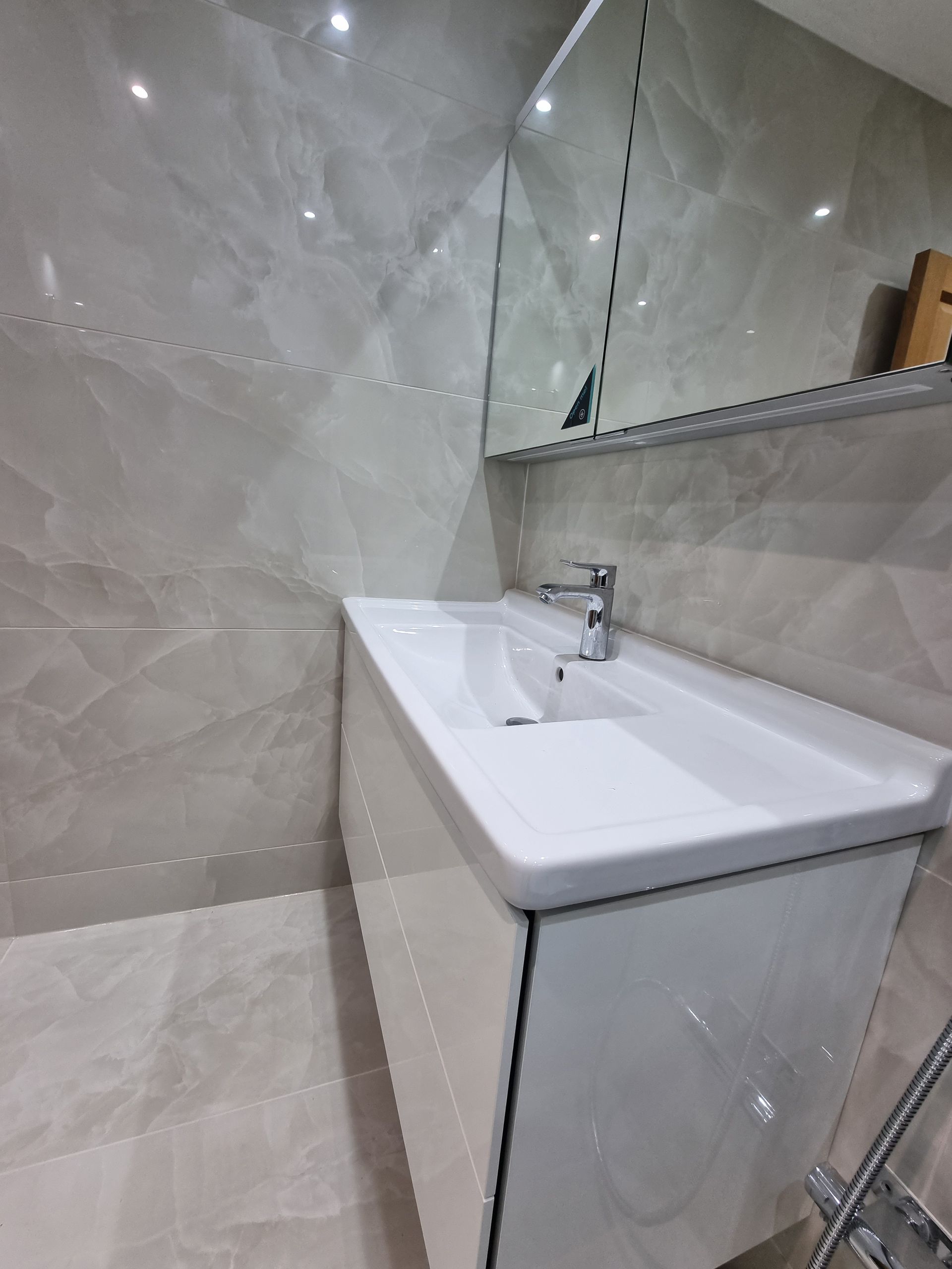 White bathroom sink with chrome faucet and mirrored cabinet, set against light gray marble tiles.
