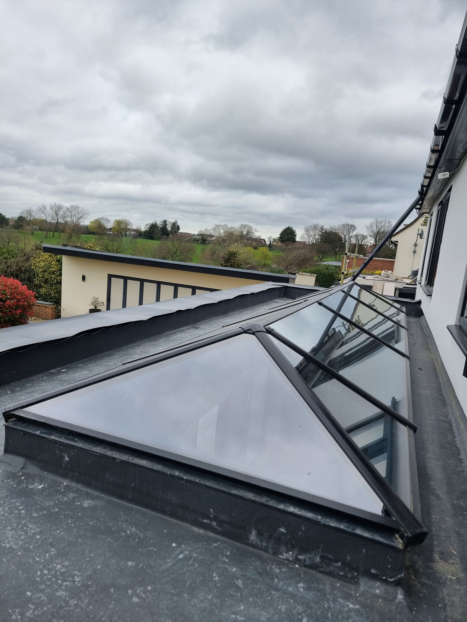 Skylight on a flat roof, black frame, glass panels, cloudy sky, residential setting.