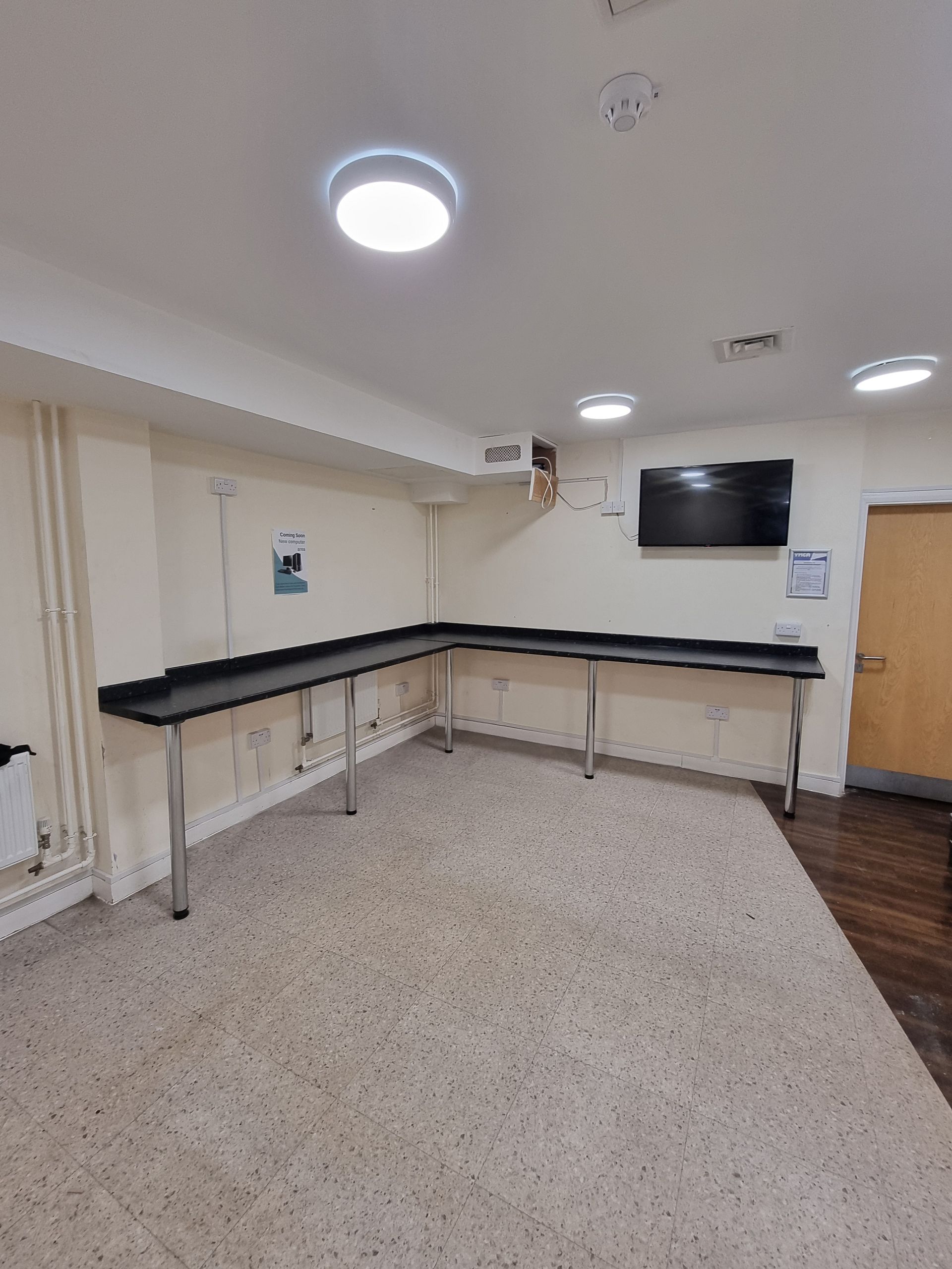 An empty room with a large L-shaped counter, a TV, and a door. Cream walls, speckled floor, and bright lighting.
