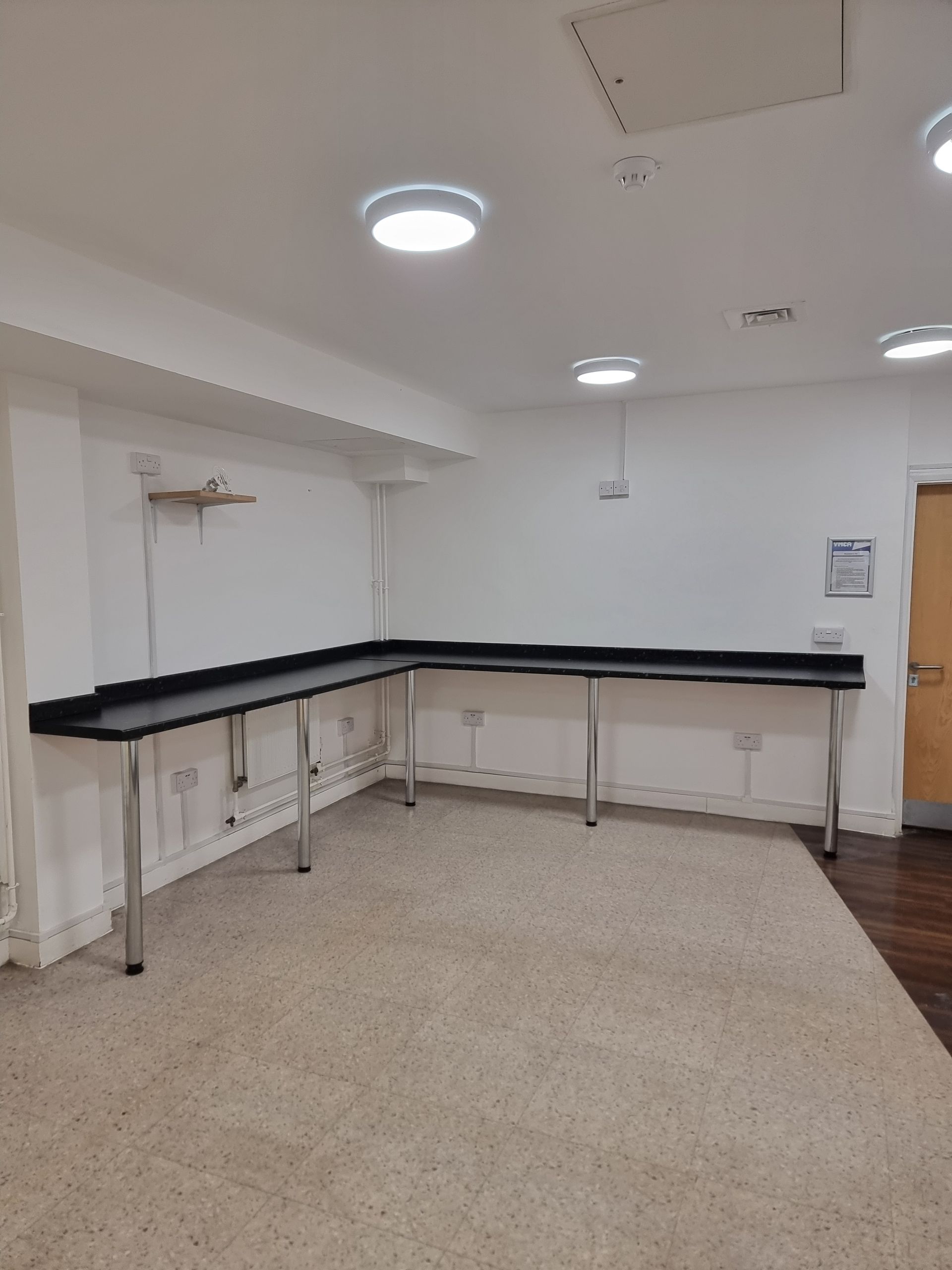 L-shaped black counter with silver legs in a white-walled room, lit by recessed ceiling lights; tan carpet.