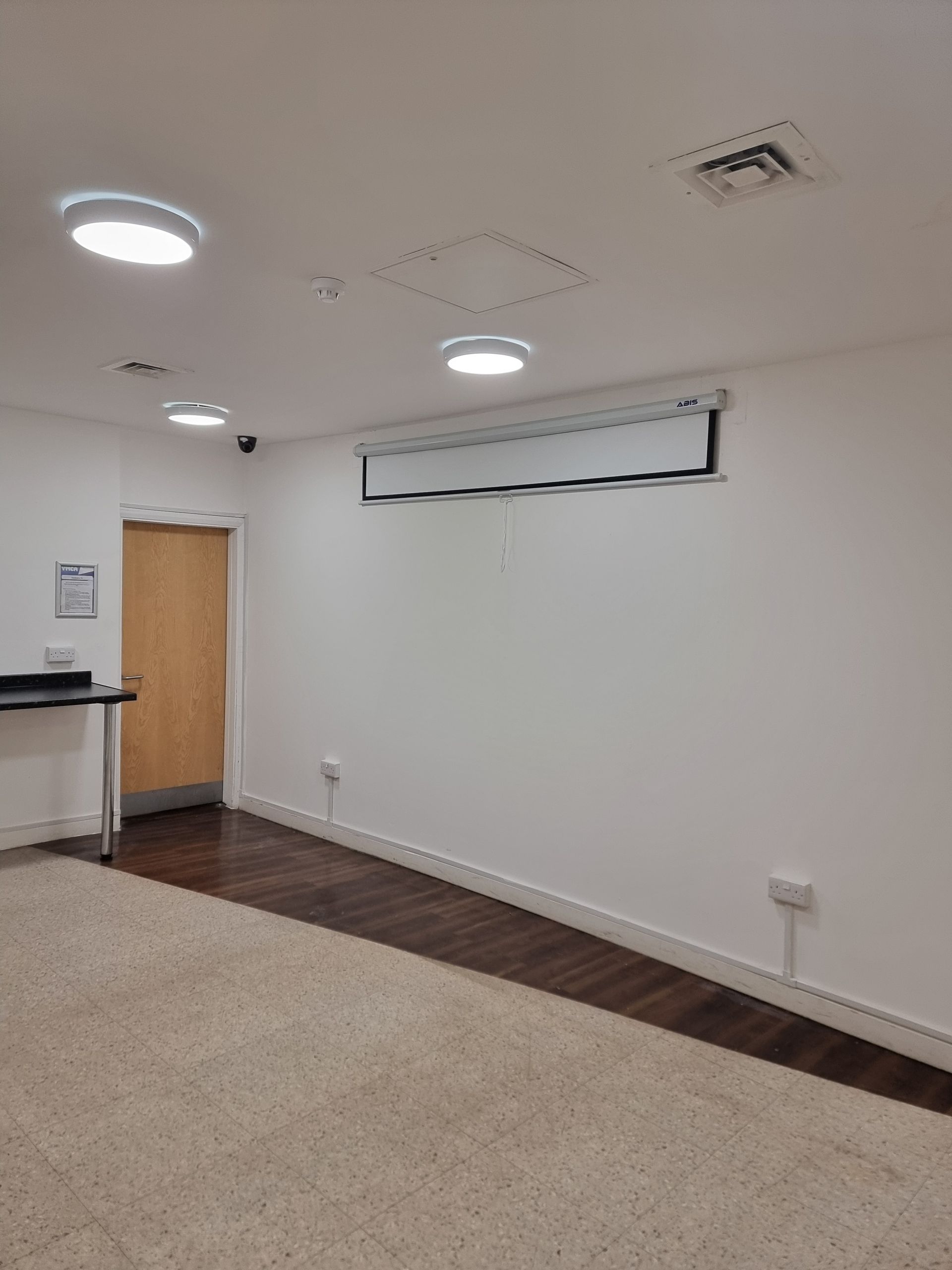 Empty room with a projector screen, wood floor, and tan carpet. The door is closed.