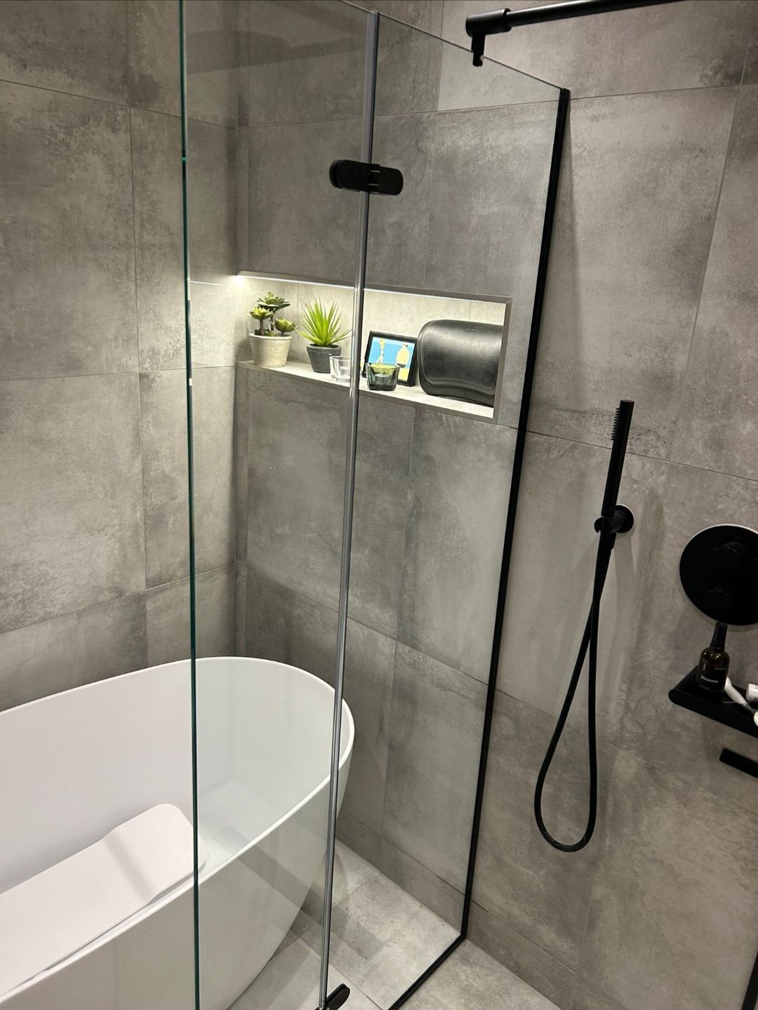 Modern bathroom with gray tiled walls, glass shower, and white bathtub. Black fixtures and a recessed shelf.