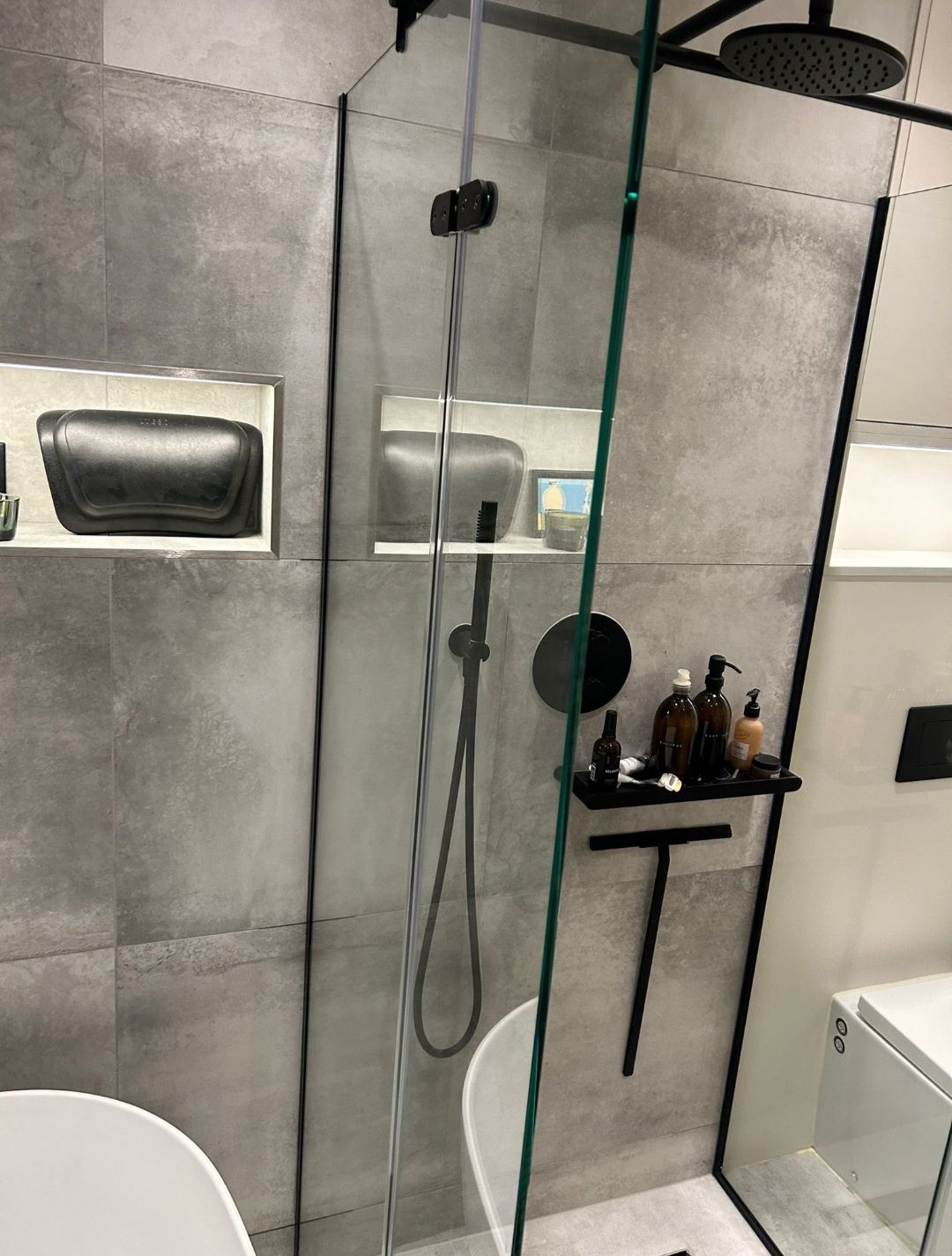 Modern bathroom with gray tiled walls, black shower fixtures, glass enclosure, and built-in shelves.