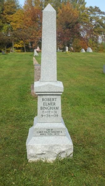 Tall granite gravestone in a grassy cemetery, with autumn trees in the background.