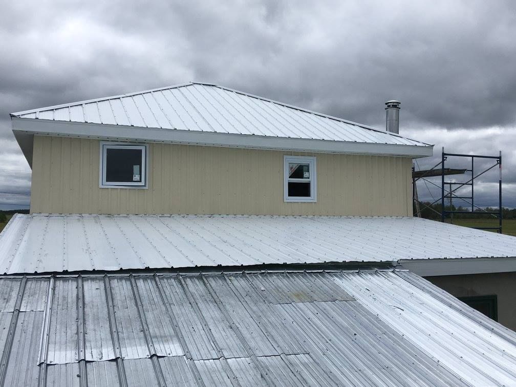 Bâtiment de deux étages avec toiture en métal blanc, bardage beige et deux fenêtres. Ciel nuageux en arrière-plan.