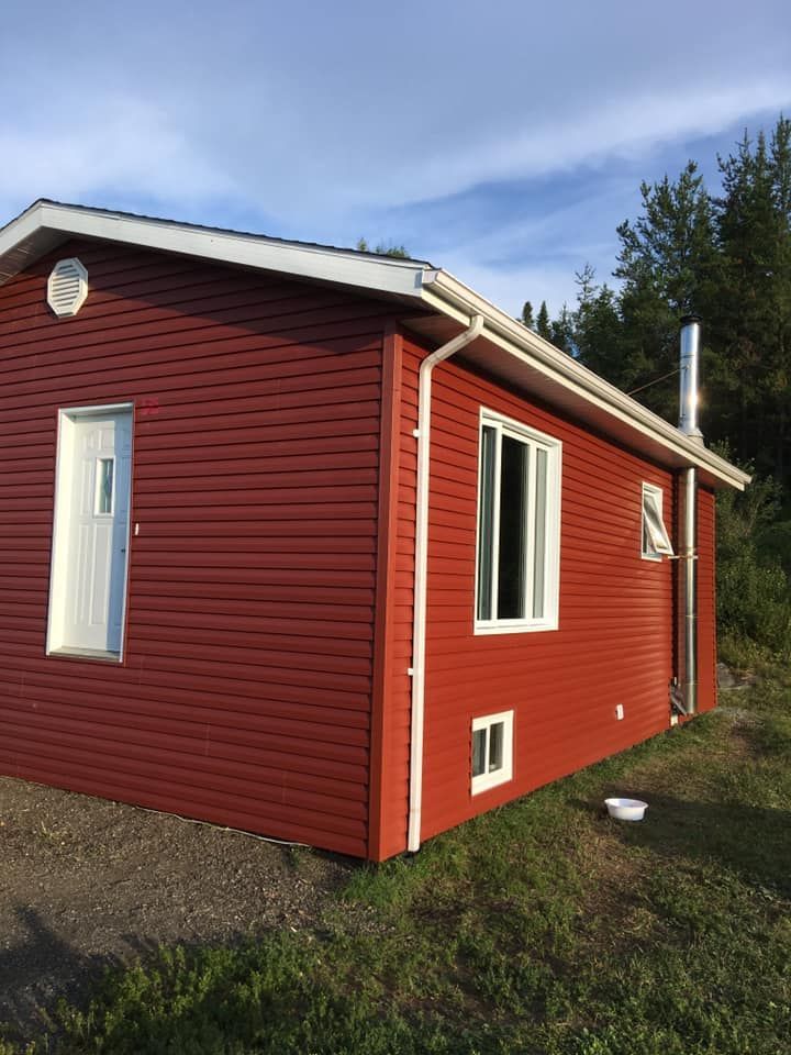 Bâtiment à façade rouge avec encadrements, porte et fenêtres blancs. Gouttières et cheminée visibles, situé sur un terrain herbeux.