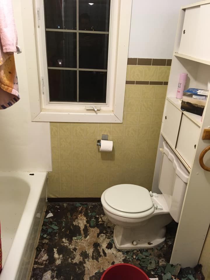 Salle de bains avec toilettes, baignoire, fenêtre, sol abîmé. Papier peint jaune, robinetterie blanche.