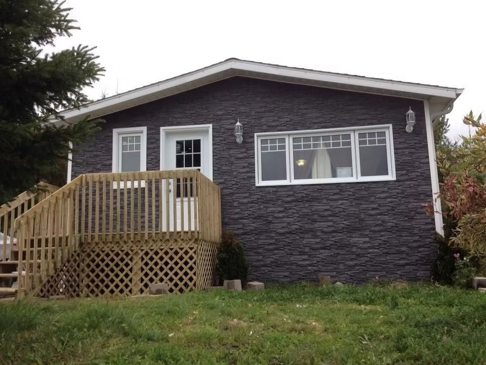 Maisonnette aux façades sombres imitant la pierre, aux boiseries blanches et à la terrasse en bois. Située sur un terrain herbeux sous un ciel nuageux.