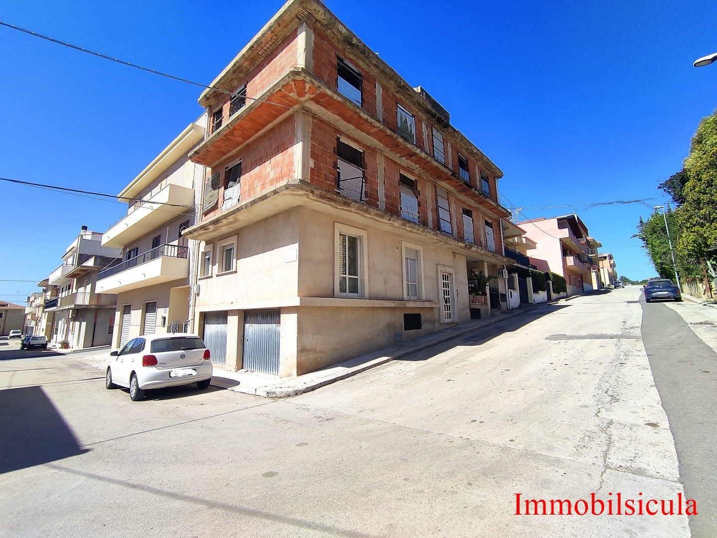 Vista d'angolo di un condominio color crema e mattoni su una strada soleggiata con auto parcheggiate e un cielo azzurro.