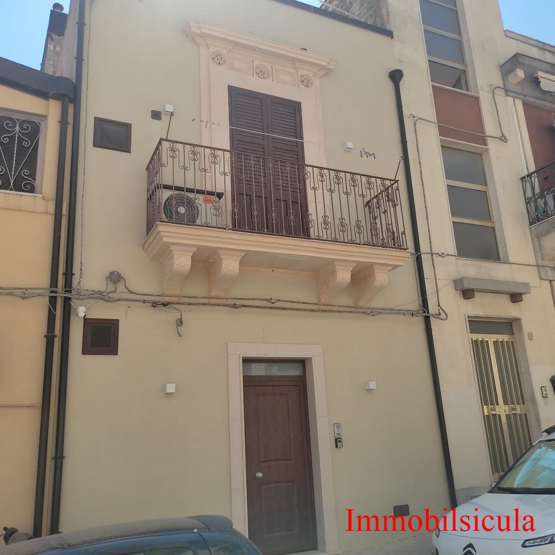 Casa a schiera a due piani color crema con balcone, porta marrone e filigrana rossa con la scritta 
