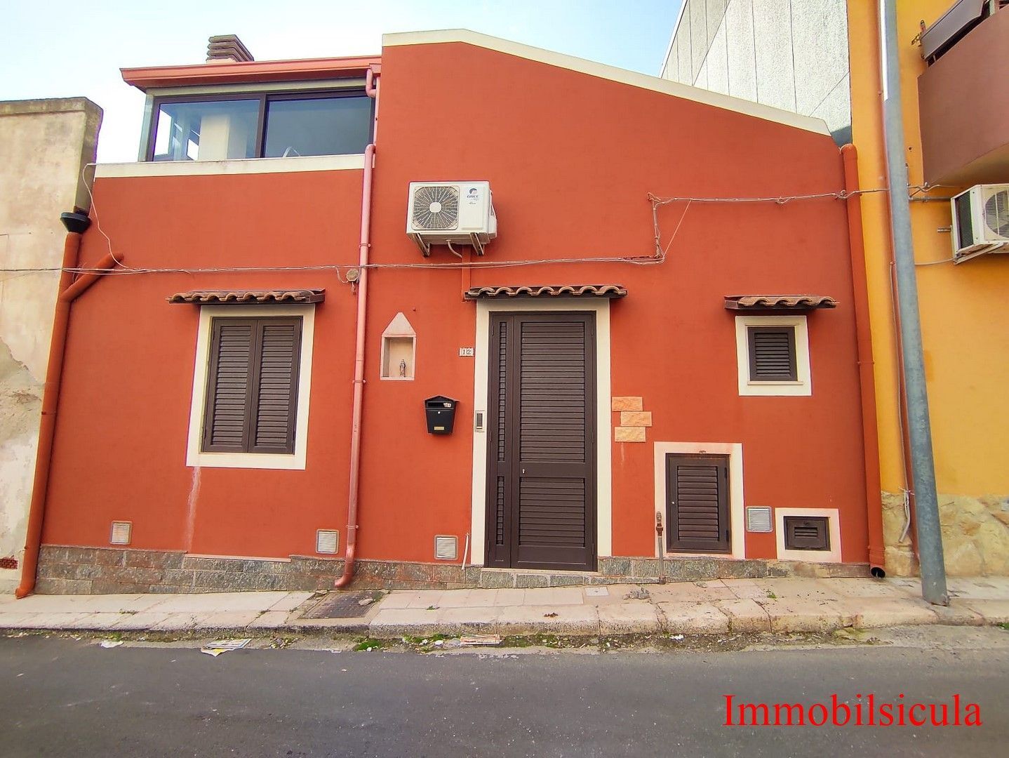 Facciata di una casa in stucco arancione con due finestre, una porta centrale e condizionatori d'aria affacciata sulla strada.
