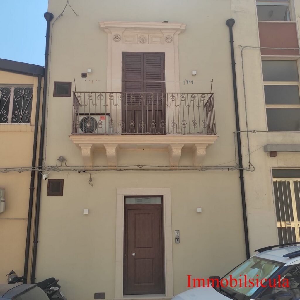 Facciata di una casa beige a due piani con balcone, porte marroni e una moto parcheggiata sul lato sinistro.