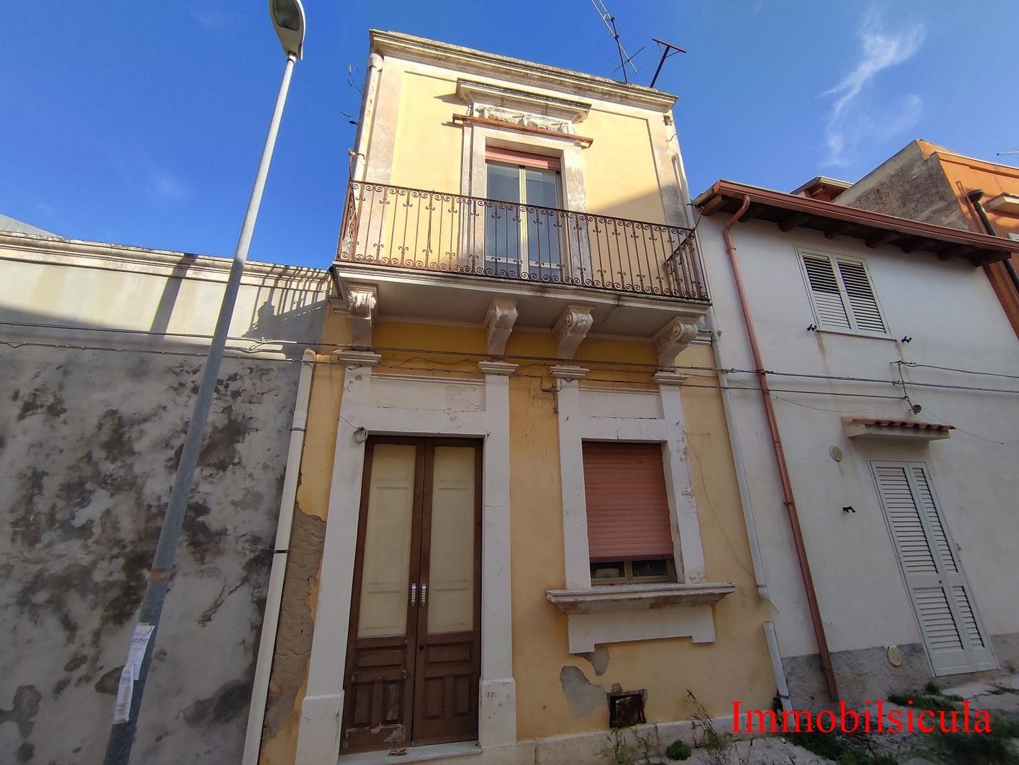 Stretta casa a schiera color giallo pastello con balcone e finestre con persiane, in una strada tranquilla sotto un cielo azzurro.