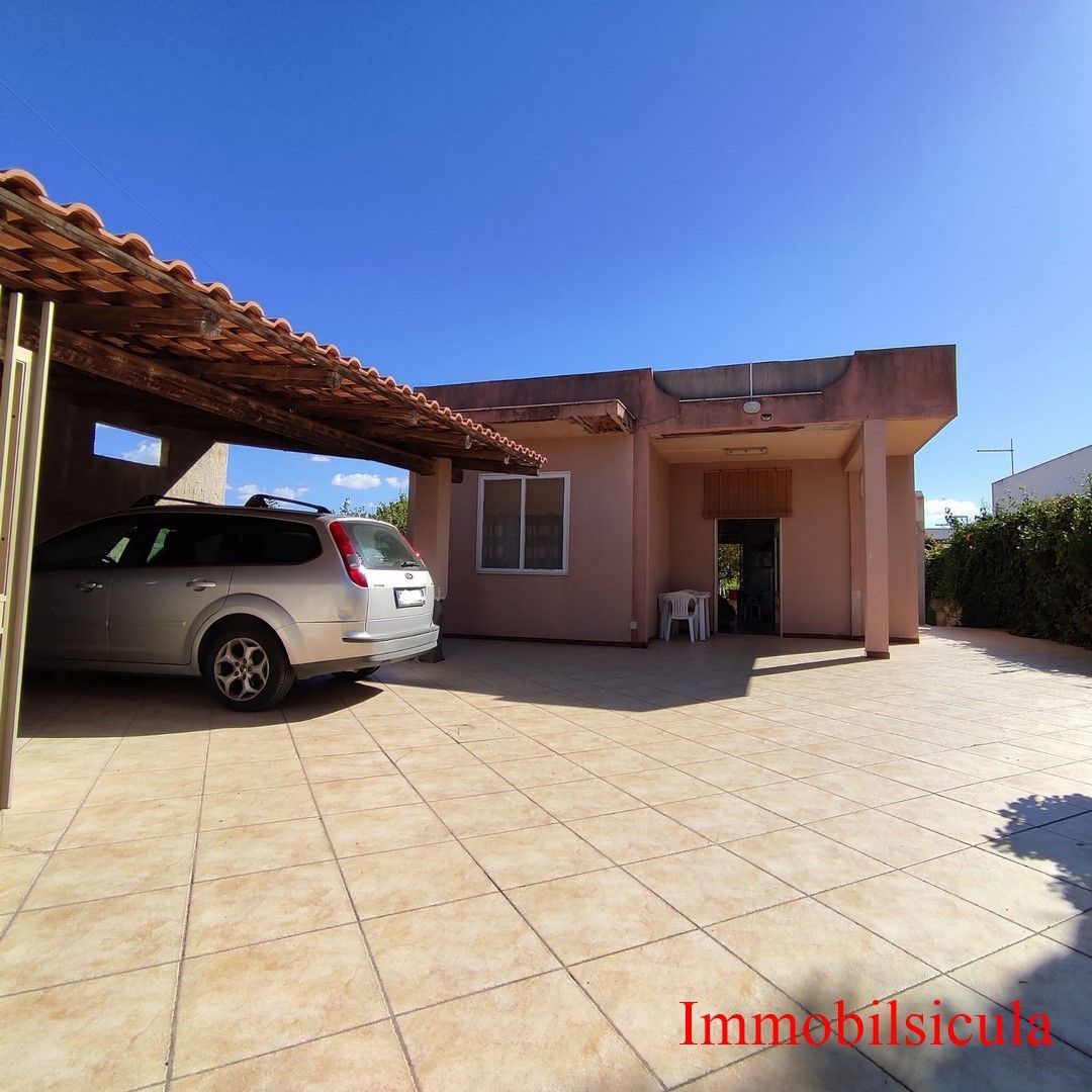 Casa a un solo piano con cortile piastrellato e un SUV argentato parcheggiato sotto una tettoia sotto un cielo azzurro