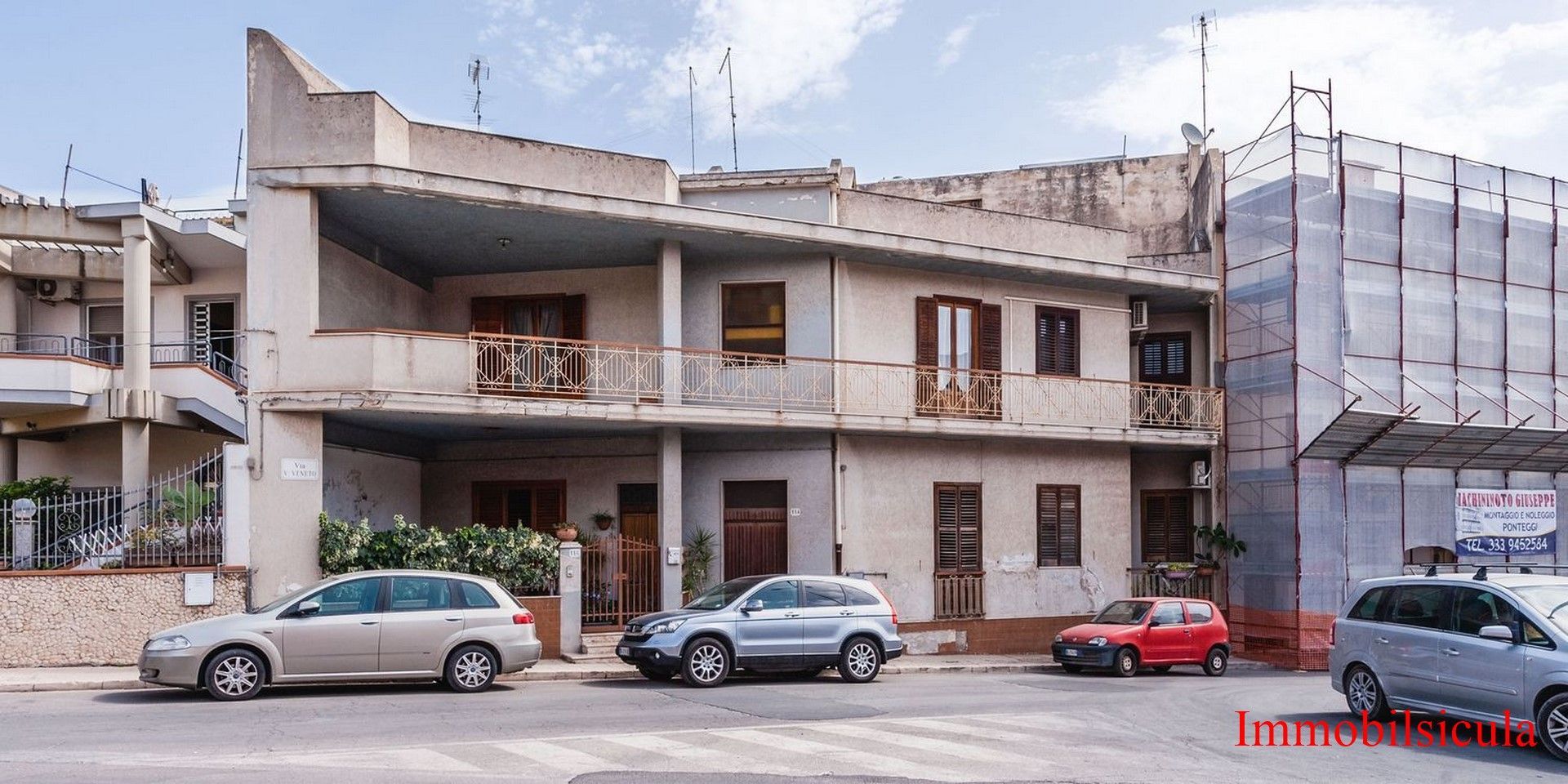 Edificio a due piani parzialmente danneggiato con auto parcheggiate su una strada, visto alla luce del giorno.