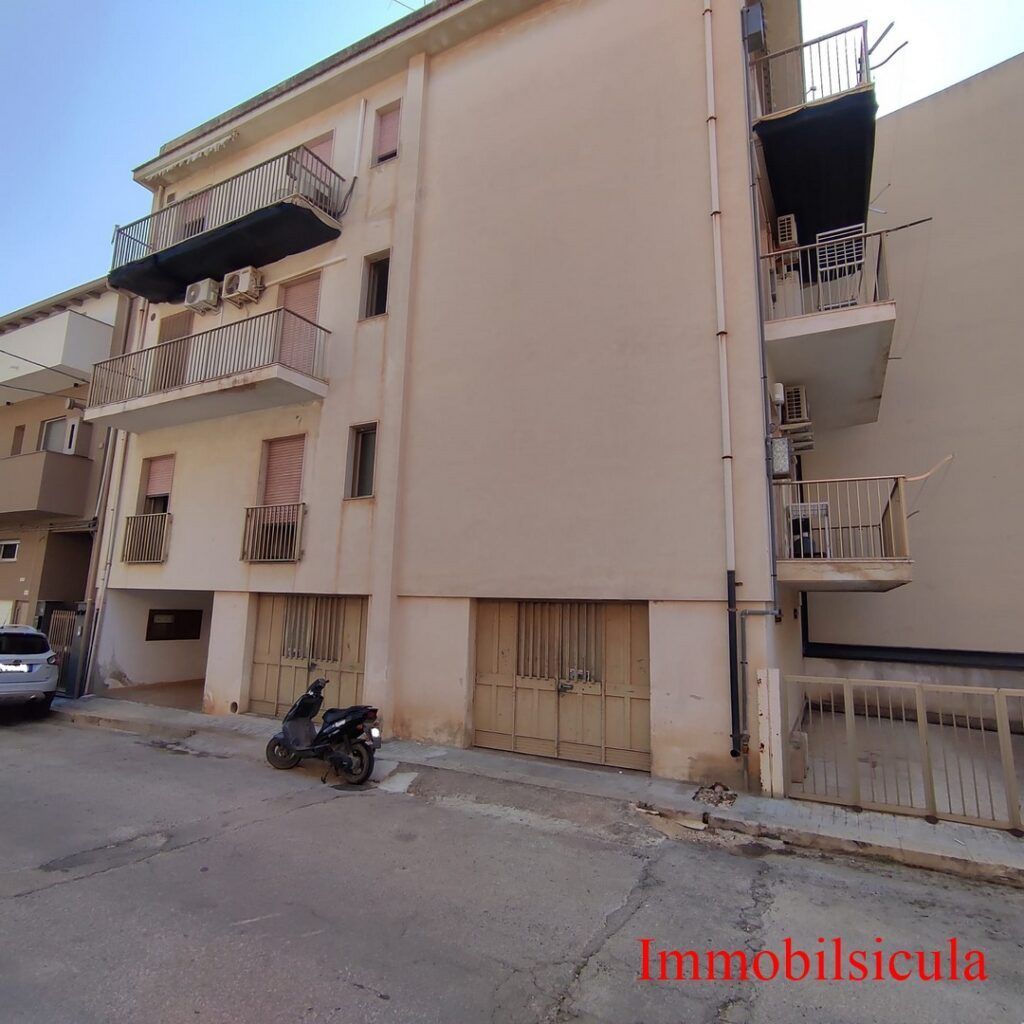 Stretta strada residenziale con palazzine beige, balconi, uno scooter parcheggiato e un'auto
