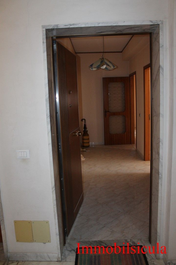 Stretto corridoio con porta aperta, pavimento piastrellato, lampada a sospensione e porte in legno.
