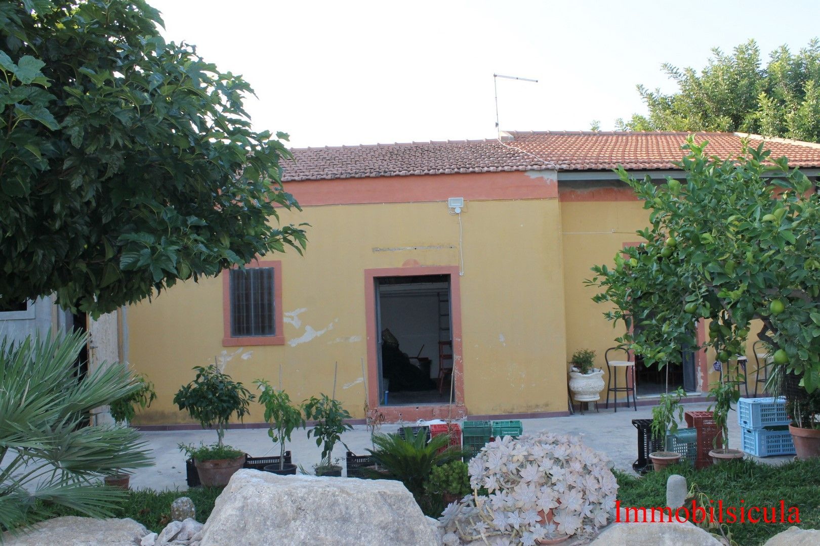 Casa gialla con tetto di tegole rosse e giardino, incorniciata da alberi e rocce in primo piano.