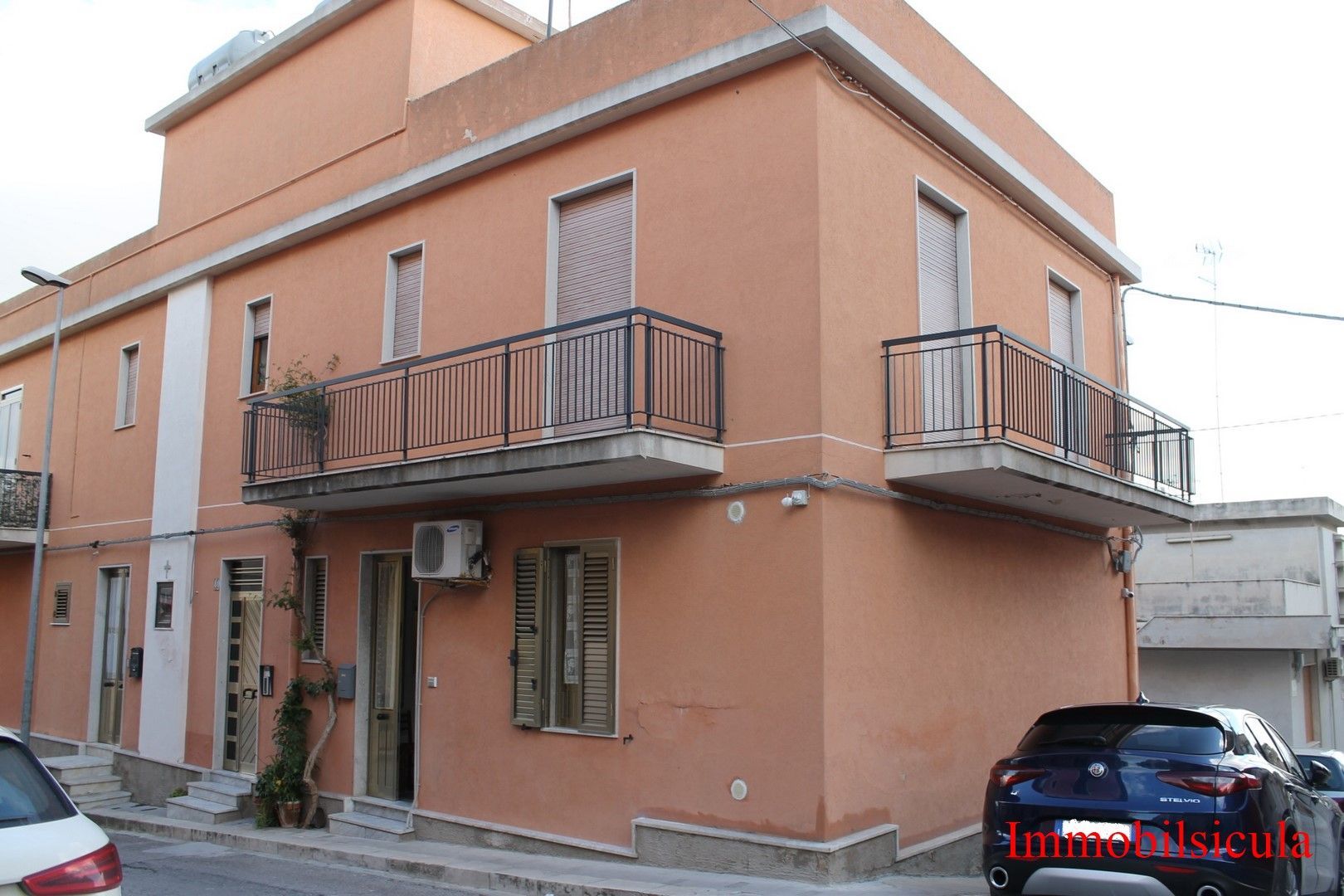 Casa arancione a due piani con balconi, situata in una strada stretta, con un'auto blu parcheggiata.
