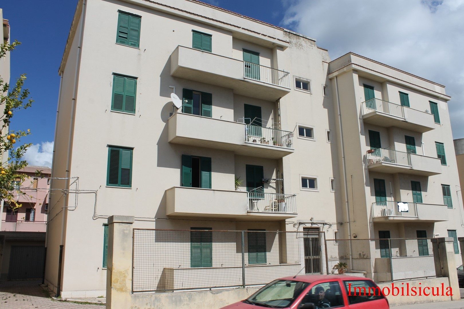 Palazzi con balconi e persiane verdi accanto a un'auto rossa parcheggiata in una strada soleggiata