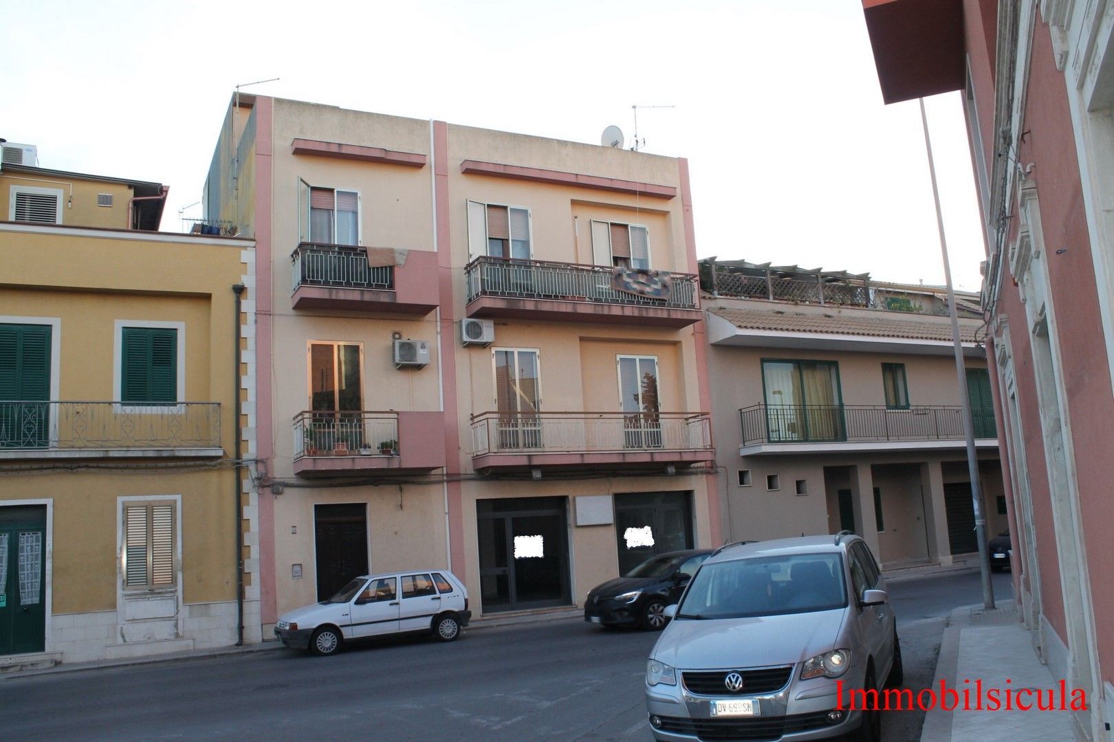 Una strada tranquilla con palazzi dai colori pastello e auto parcheggiate sotto un cielo nuvoloso.