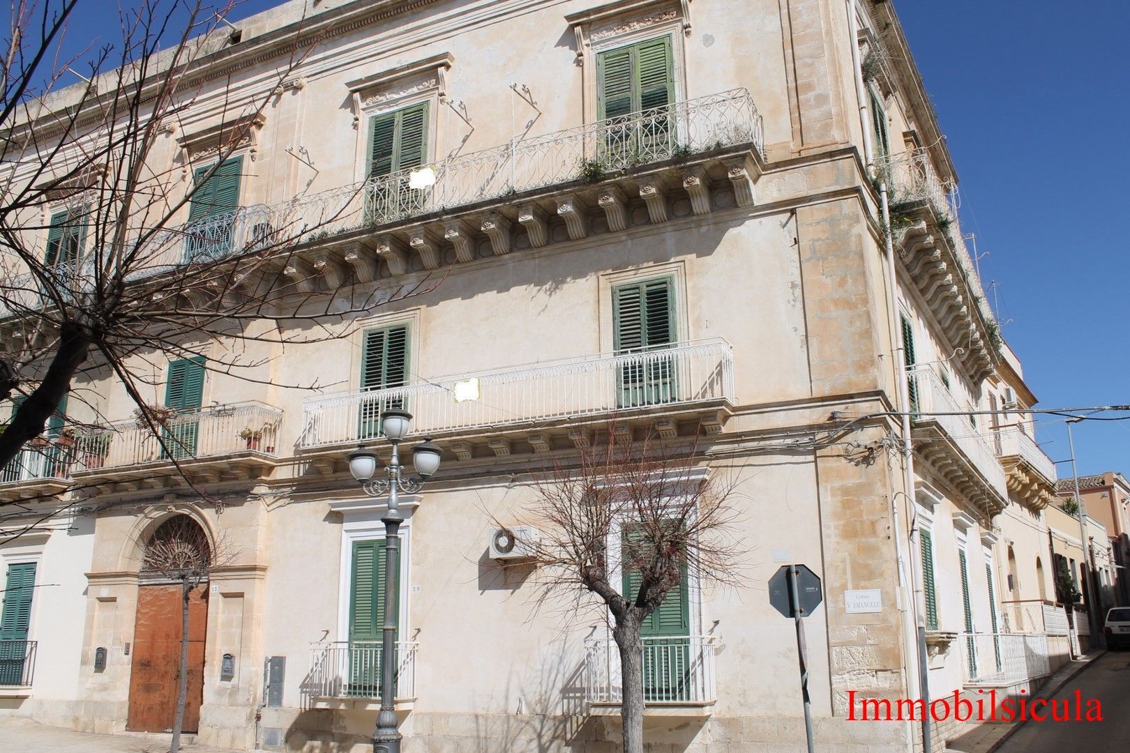 Vecchio palazzo beige con persiane e balconi verdi, affacciato su una strada soleggiata.