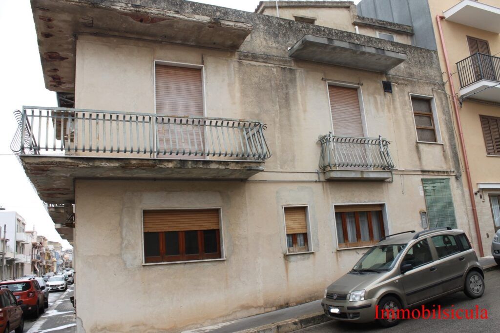 Vecchia casa d'angolo beige con balconi su una strada in pendenza, auto parcheggiata davanti.
