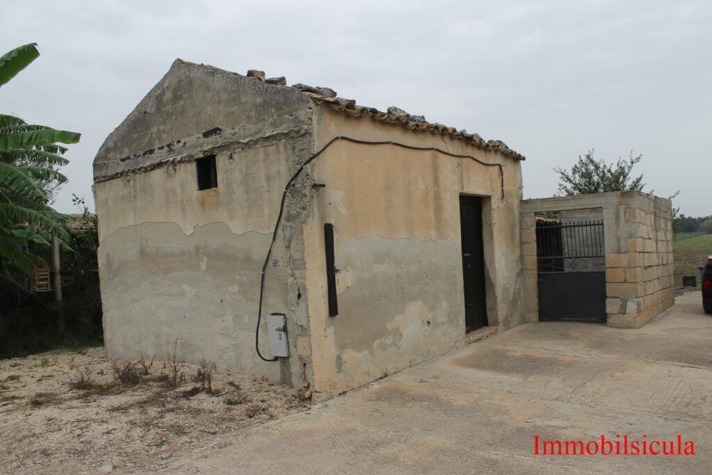 Una piccola casa beige, consumata dal tempo, con tetto di paglia e porta aperta, in un cortile di campagna.