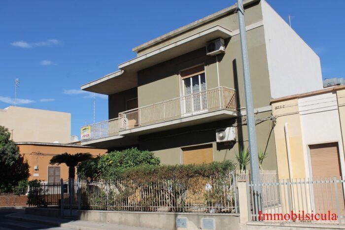 Casa beige a due piani con balconi, una recinzione e un cielo azzurro.