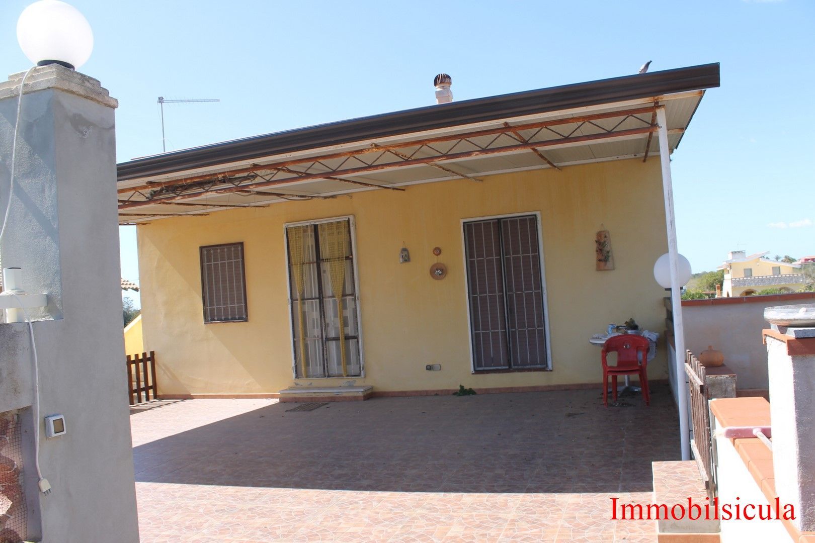 Casa gialla con portico coperto, porte in metallo e patio anteriore piastrellato in piena luce del sole